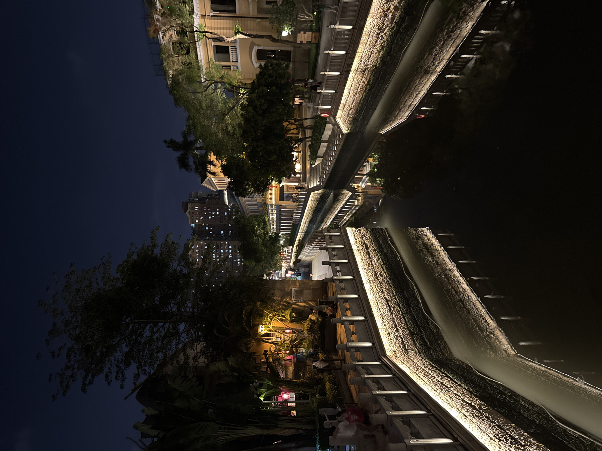 This is an outdoor nighttime scene in what appears to be a park or pedestrian area in the Liwan District of China. The image seems to be taken at ground level. A waterway reflects the buildings and light features along the sides.
The buildings and the river banks are illuminated with artificial lights. Some people are present along the walkway, some seated and some standing. The sky is dark blue. The scene includes trees and other vegetation mixed in with the buildings.