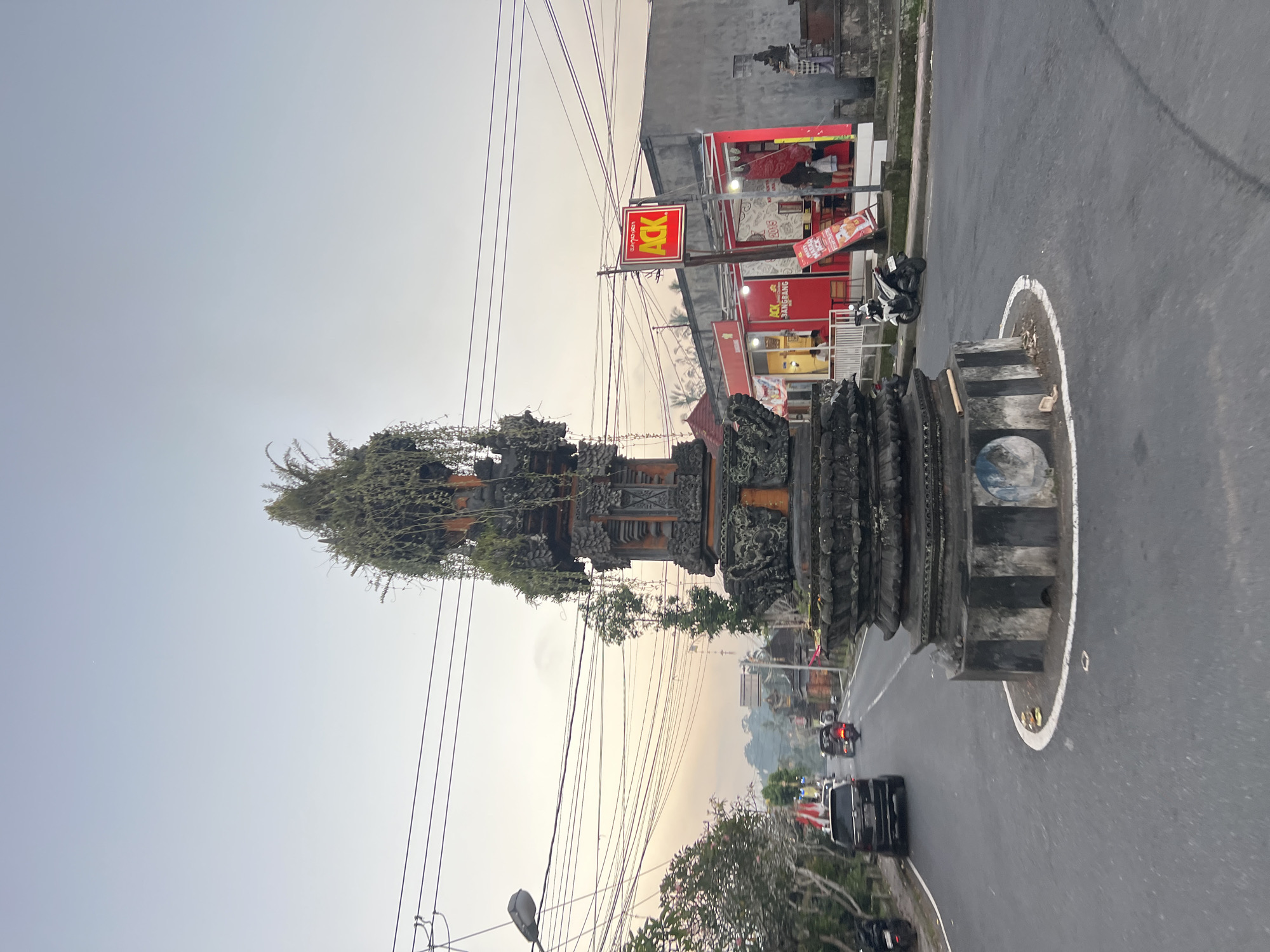In Tembuku, Indonesia, a street-level, medium-shot photograph captures a traffic circle dominated by a traditional Balinese temple-like structure. The structure, made of dark stone with intricate carvings, is topped with lush green foliage, creating a unique blend of natural and man-made elements. To the right of the traffic circle, a red and white building with a prominent "ACK" sign stands, appearing to be a commercial establishment, possibly a restaurant. Several motorcycles and a car are visible on the asphalt road, indicating moderate traffic. The sky is overcast with a soft, diffused light, suggesting it is either early morning or late afternoon. The environment appears to be a mixed urban and rural setting, with trees lining the road and power lines crisscrossing the sky. The overall scene conveys a sense of daily life in a Southeast Asian town, with its characteristic blend of cultural heritage and modern infrastructure.