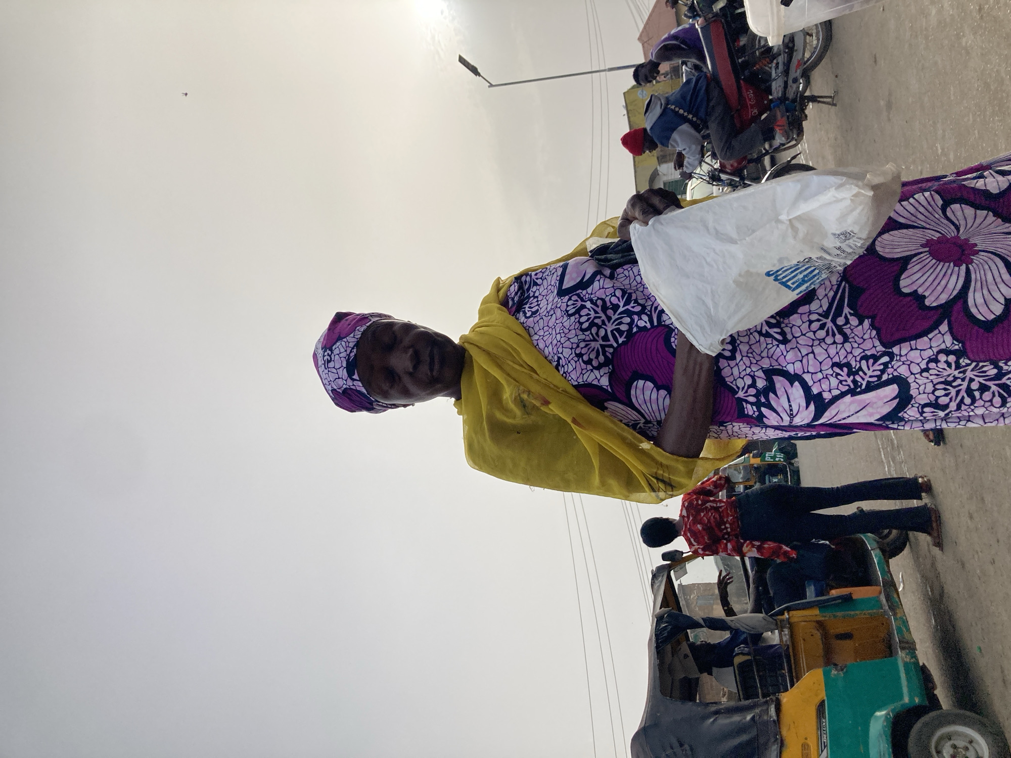 The image captures an outdoor scene, likely a busy street or market area in Gwong, Nigeria, under a bright, somewhat hazy sky. The primary subject is a woman standing in the foreground, slightly to the right of the center. She wears a vibrant purple and white patterned dress or wrapper, a matching head covering, and a yellow scarf or shawl draped over her shoulders. She holds a white plastic bag in her left hand and looks towards the viewer with a calm expression.
In the mid-ground, to the right, a person in a red patterned shirt and dark trousers stands beside a green and yellow tricycle (keke napep), with another individual visible seated inside. Several motorcycles and more people are present in the background, suggesting a lively environment. A street light pole and thin power lines are visible against the diffused light of the sky. The ground appears to be paved. Part of the word "GLORY" is legible on the white plastic bag the woman is carrying. The bright conditions indicate it is daytime.