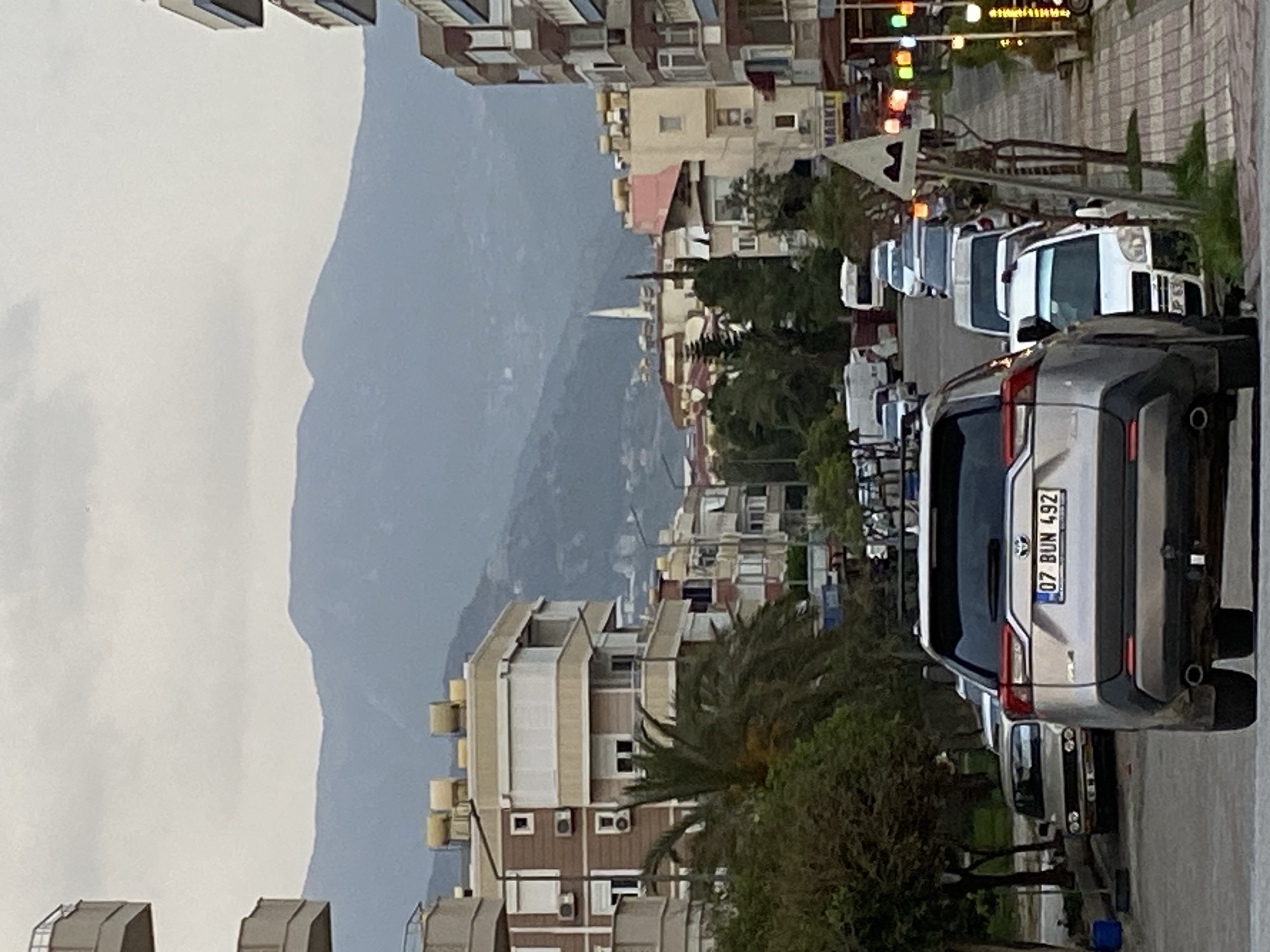 The image shows a street in Alanya, Turkey, with buildings lining both sides of the road. In the background are mountains shrouded in mist. In the foreground, several cars are parked along the street, with a silver Toyota RAV4 occupying a prominent position, its license plate reading "07 BUN 492." Trees and other vegetation grow along the roadside. The weather appears overcast. Traffic lights are visible in the distance. The architecture includes a mix of residential and commercial buildings. A minaret can be seen between the buildings.