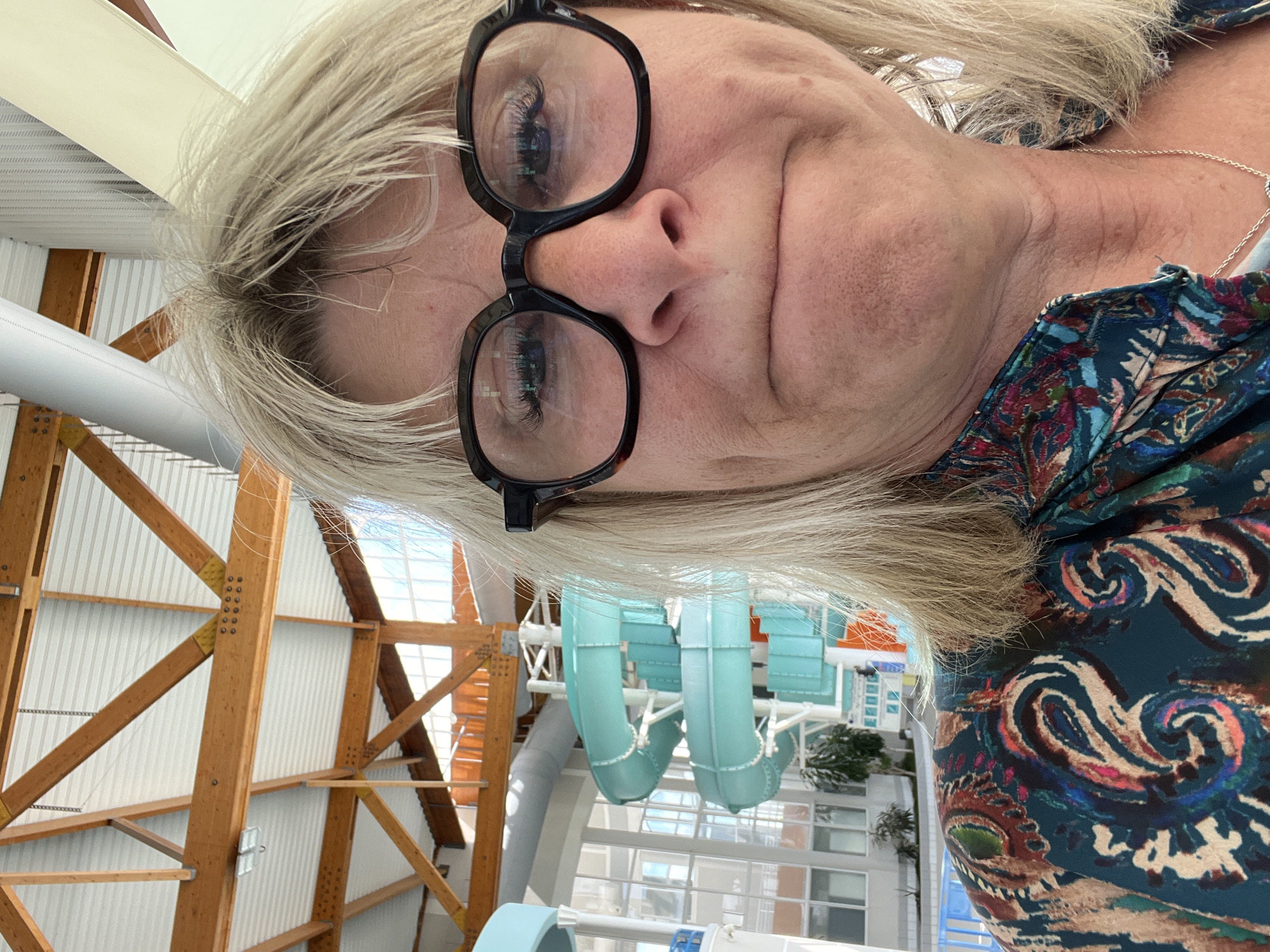 This close-up image captures a woman at an indoor aquatic center, likely a water park. The woman, with blonde hair and black-framed glasses, is positioned towards the left of the frame, looking towards the camera with a neutral to slightly contemplative expression. Her face is in focus, and reflections of her eyes and the environment are visible in her glasses. She is wearing a colorful top with a paisley-like pattern in hues of teal, brown, and red, and a thin silver chain is visible around her neck.
The background reveals the spacious interior of the aquatic facility. Large wooden structural beams, forming a complex framework, span across the upper and central parts of the scene. Behind these beams are white corrugated panels, likely part of the ceiling or upper walls. Sections of light blue water slides, typical of a water park, are visible in the mid-ground, curving downwards. Large windows in the background indicate it is daytime, allowing natural light to illuminate the facility. The scene suggests a relaxed atmosphere in a recreational setting. No discernible text is visible on any objects.
