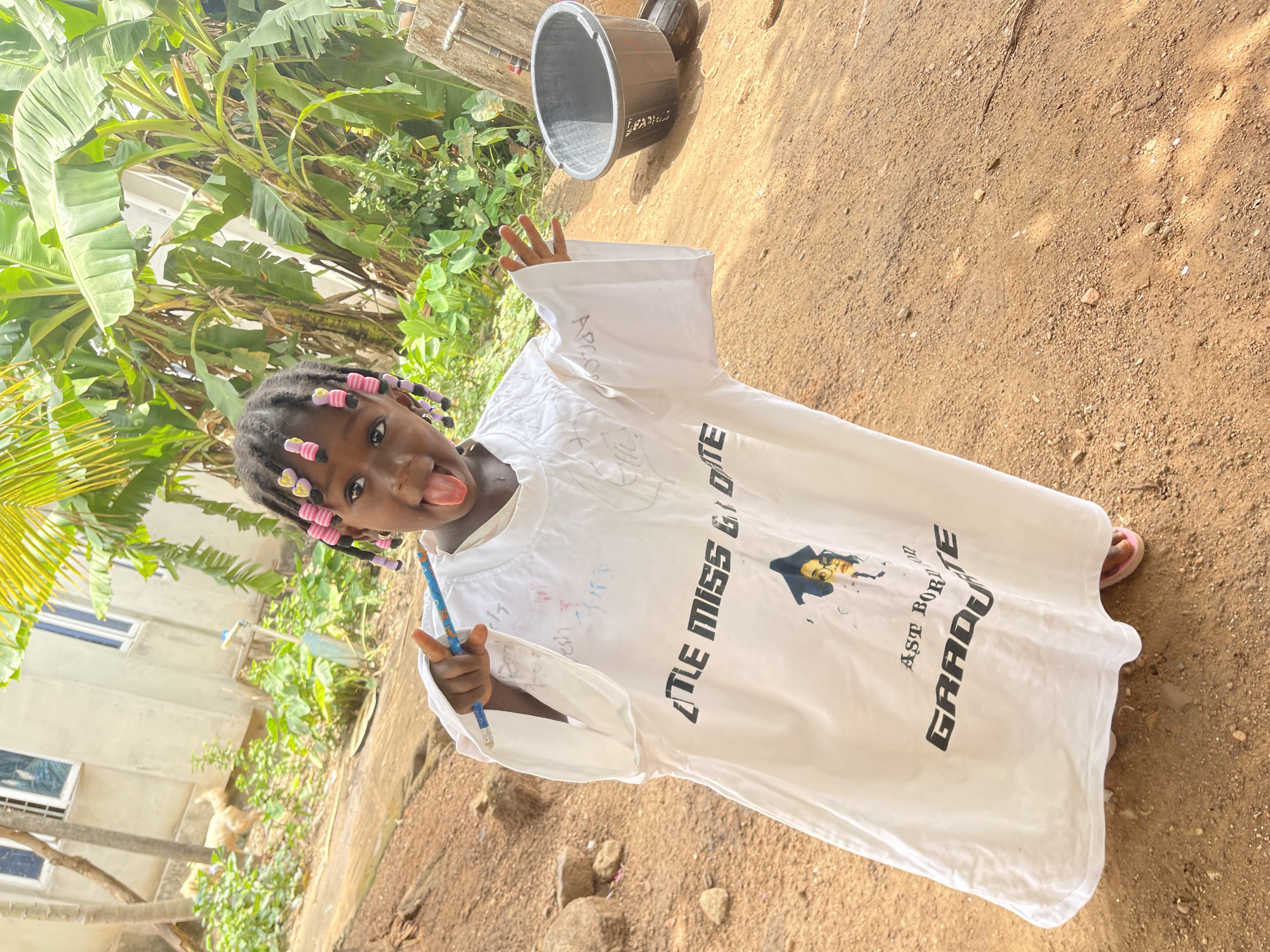 A young Black child, likely a girl, is captured in a playful pose outdoors, sticking her tongue out towards the camera. She has dark braided hair adorned with colorful pink, purple, and yellow beads. In her right hand, she holds a blue pencil, pointing it downwards.
She is wearing an oversized white t-shirt that reaches past her knees. The t-shirt features prominent black text across the chest: "LITTLE MISS GRADUATE," and further down, "GRADUATE" with smaller text "AST BORN" above it. A graphic of a person wearing a graduation cap and glasses is also printed on the shirt. Faint, hand-drawn scribbles or writing are visible on the white fabric. Her feet, clad in light-colored sandals, are partially visible beneath the long shirt.
The child stands on a surface of dry, reddish-brown dirt mixed with small stones and patches of light-green vegetation. The background is rich with tropical foliage, including large, vibrant green banana leaves on the left side, and other lush plants. A light-colored building with windows is partially visible behind the dense greenery on the left. In the mid-ground, a light-feathered fowl, possibly a duck or chicken, is seen near some plants and another building structure. A dark-colored bucket rests on the ground in the upper background.
The scene is brightly illuminated by natural daylight, indicating a sunny day. The child's cheerful and mischievous expression, combined with the "GRADUATE" themed shirt, suggests a moment of celebration or pride. The environment, characterized by tropical flora, dry ground, and simple structures, is consistent with a rural or semi-urban setting in Akwanga, Nigeria.