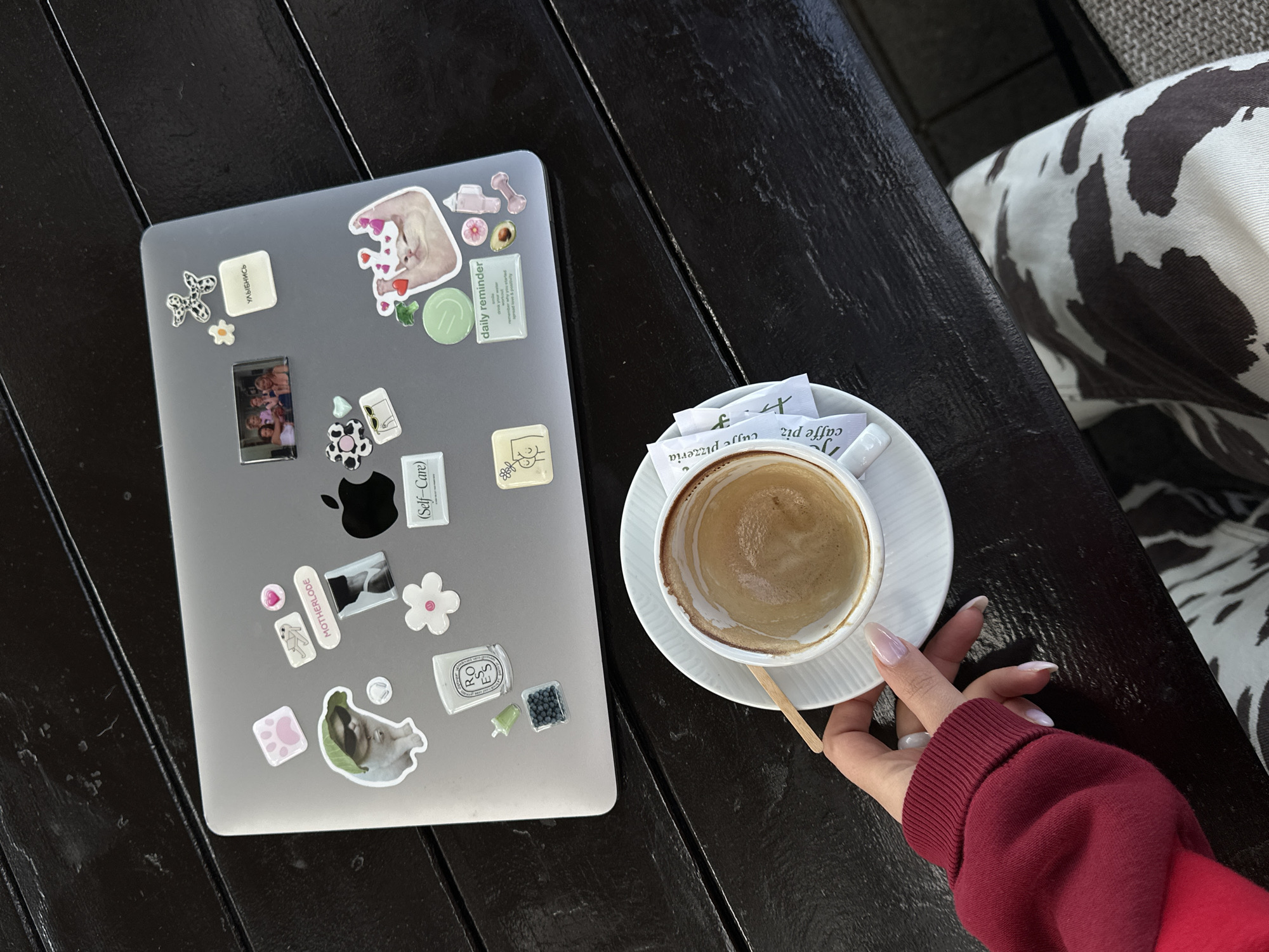 The image shows a laptop covered in stickers on a dark wooden table. To the right of the laptop is a white cup of coffee on a matching saucer. A small wooden stirrer rests in the saucer. Packets of sugar are also present on the saucer. A person's hand, wearing a red sweater, is holding the saucer. Part of a patterned seat is visible on the right. The image suggests a cafe setting, possibly in Šušanj, Montenegro, based on the location.