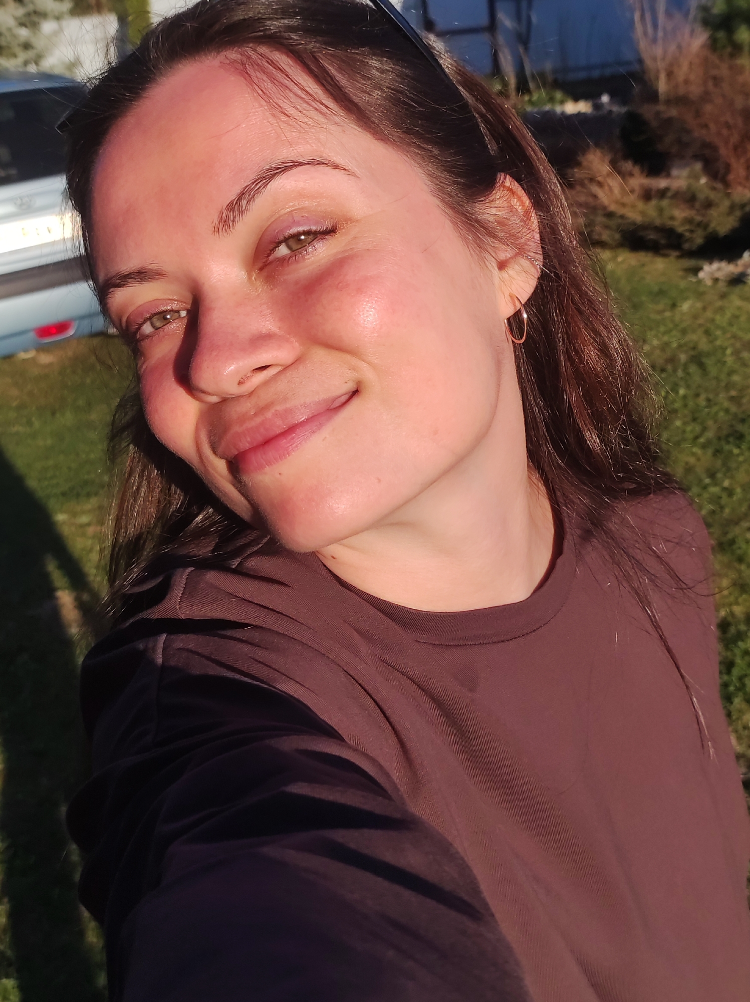 A woman with brown hair and a brown shirt is smiling at the camera. She is wearing gold hoop earrings. A vehicle is partially visible in the background. The setting appears to be outdoors, possibly a yard or field with grass. The image is lit by sunlight, with shadows and highlights on the woman's face and shirt. There is no indication that the image was taken in the city of Фоки, Russia.