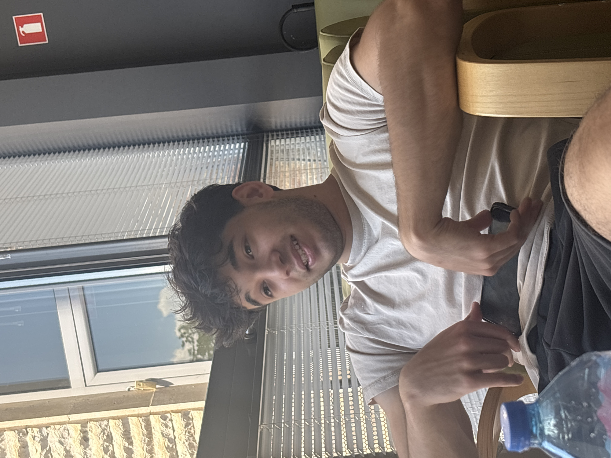 The image shows a young man sitting indoors, rotated 90 degrees counter-clockwise. He has dark, curly hair and a light beard, wearing a light-colored t-shirt. He is looking at the camera with a slight smile, suggesting a relaxed mood. His hands are visible, one holding what appears to be a smartphone.
The setting appears to be a modern indoor space, possibly a cafe, office, or communal area, characterized by large windows with horizontal blinds that let in bright daylight. Outside the window, a brick-like building facade and glimpses of trees and sky with clouds are visible, indicating a daytime scene with sunny weather. A wooden table or counter is in front of the man, and a dark-colored chair or seat back is behind him. In the bottom right foreground, a plastic water bottle with a blue cap is partially visible.
In the upper left corner of the image, on a dark wall or ceiling, there is a red square sign with a white graphic of a fire extinguisher, indicating safety equipment. The presence of such a sign, combined with the style of windows and interior, aligns with a public or commercial building in a location like Blagoevgrad, Bulgaria. The man's attire and relaxed pose suggest a casual environment.