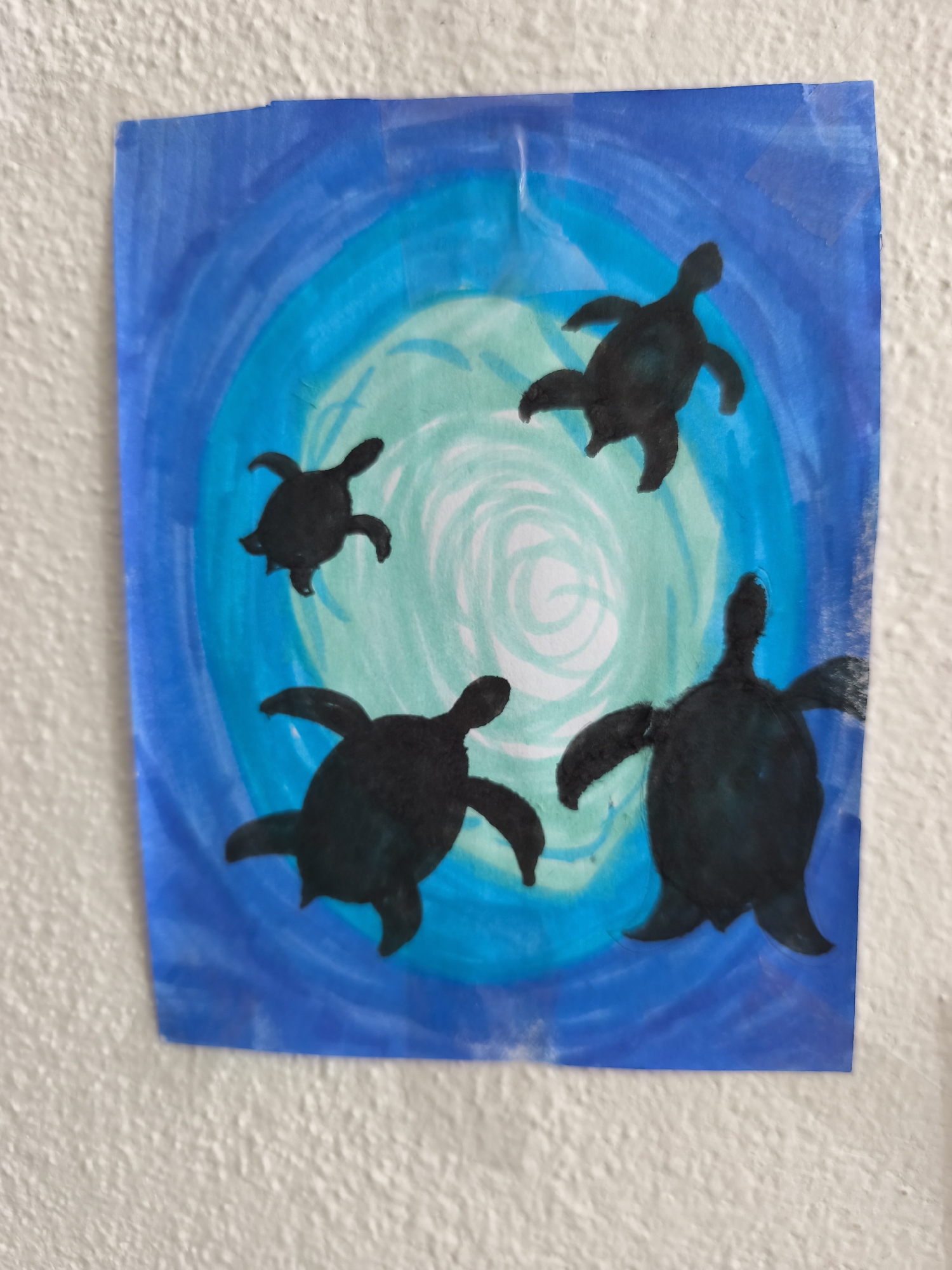 The image shows a child's drawing of four black silhouetted sea turtles swimming in blue and green watercolor. The drawing is taped to a textured, off-white wall. The turtles are in various positions, with one larger turtle in the foreground on the right, and three smaller turtles to the left and top of the frame. The background depicts concentric circles of blue and green, suggesting water or ripples. The drawing appears to be on a piece of paper that is slightly larger than the artwork itself, with some of the tape visible at the top edge. The overall impression is of a simple, colorful artwork. The location of this artwork is unknown and cannot be determined from the image provided.