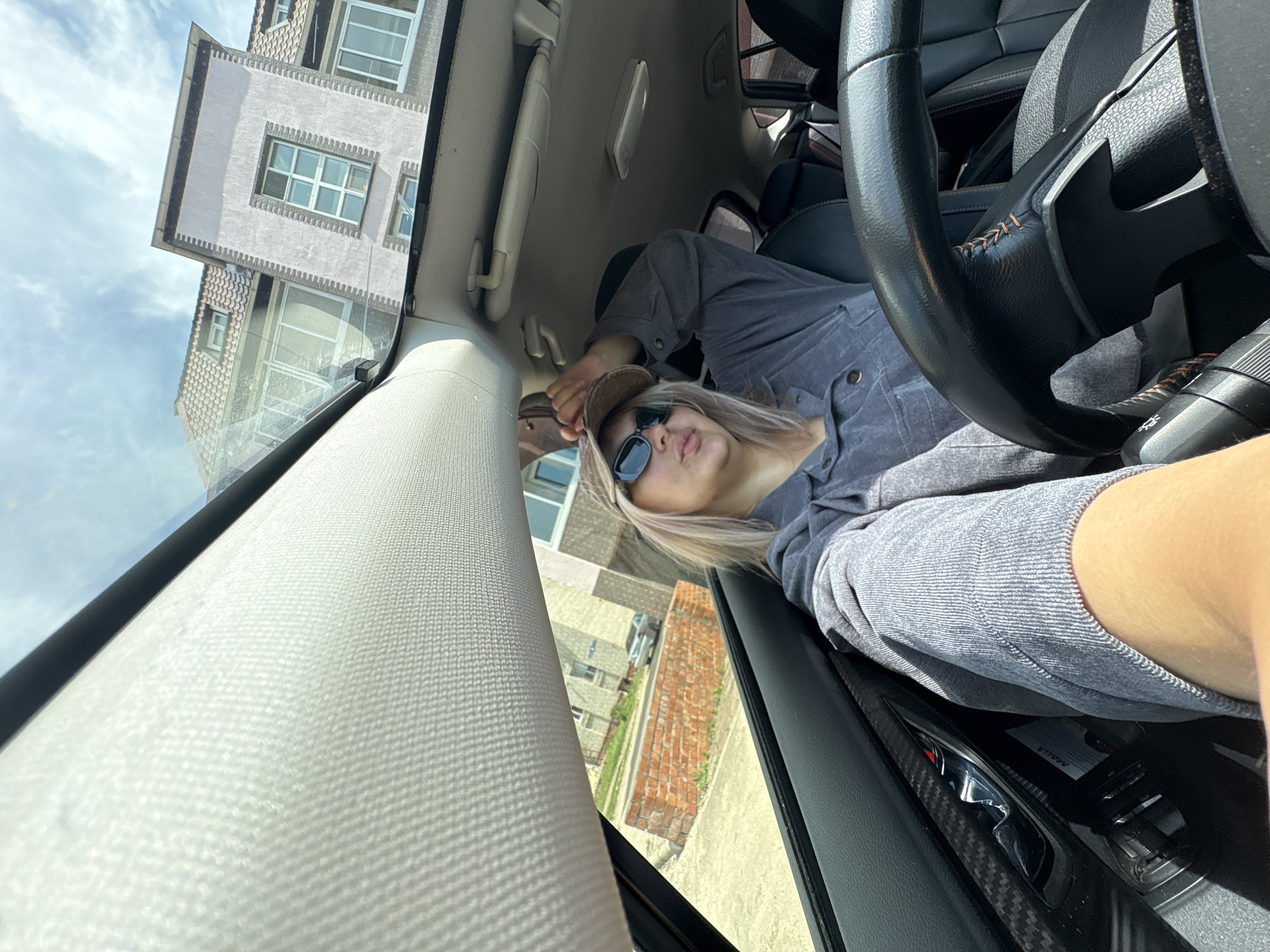 A woman with blonde hair, wearing sunglasses and a baseball cap, sits in the driver's seat of a car. She is holding a baseball cap in her right hand and touching her head with it. The car's interior, including the steering wheel and dashboard, is visible. Through the car's windows and sunroof, buildings and a brick wall are visible, suggesting an urban setting. The sky is cloudy. This image was captured in the city of Birobidzhan, Russia.