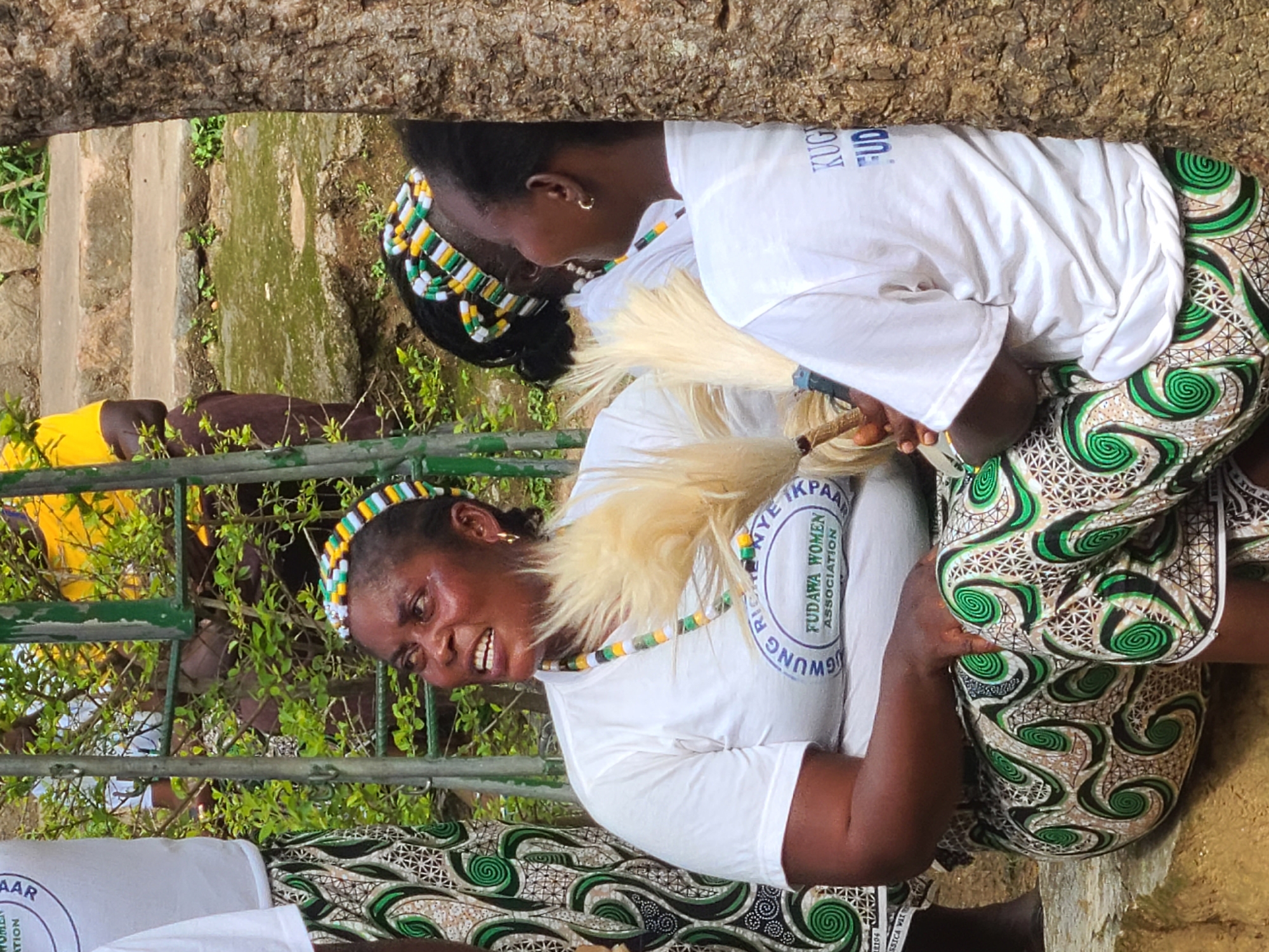 In an outdoor setting, likely in Jos, Nigeria, a group of women dressed in traditional attire are gathered. The foreground features a woman wearing a white t-shirt with a circular logo that reads "FUDOWA WOMEN ASSOCIATION" and a colorful beaded headband. She is seated and holding what appears to be a fan made of straw. Next to her, another woman is also seated, wearing a similar beaded headband and white t-shirt, with her face partially obscured. Behind them, a person in a yellow shirt is visible. The environment includes a metal fence, lush green foliage, and a rough, textured tree trunk on the right side of the frame. The ground appears to be uneven and somewhat damp. The overall impression is a communal gathering or event, possibly a cultural celebration or meeting.