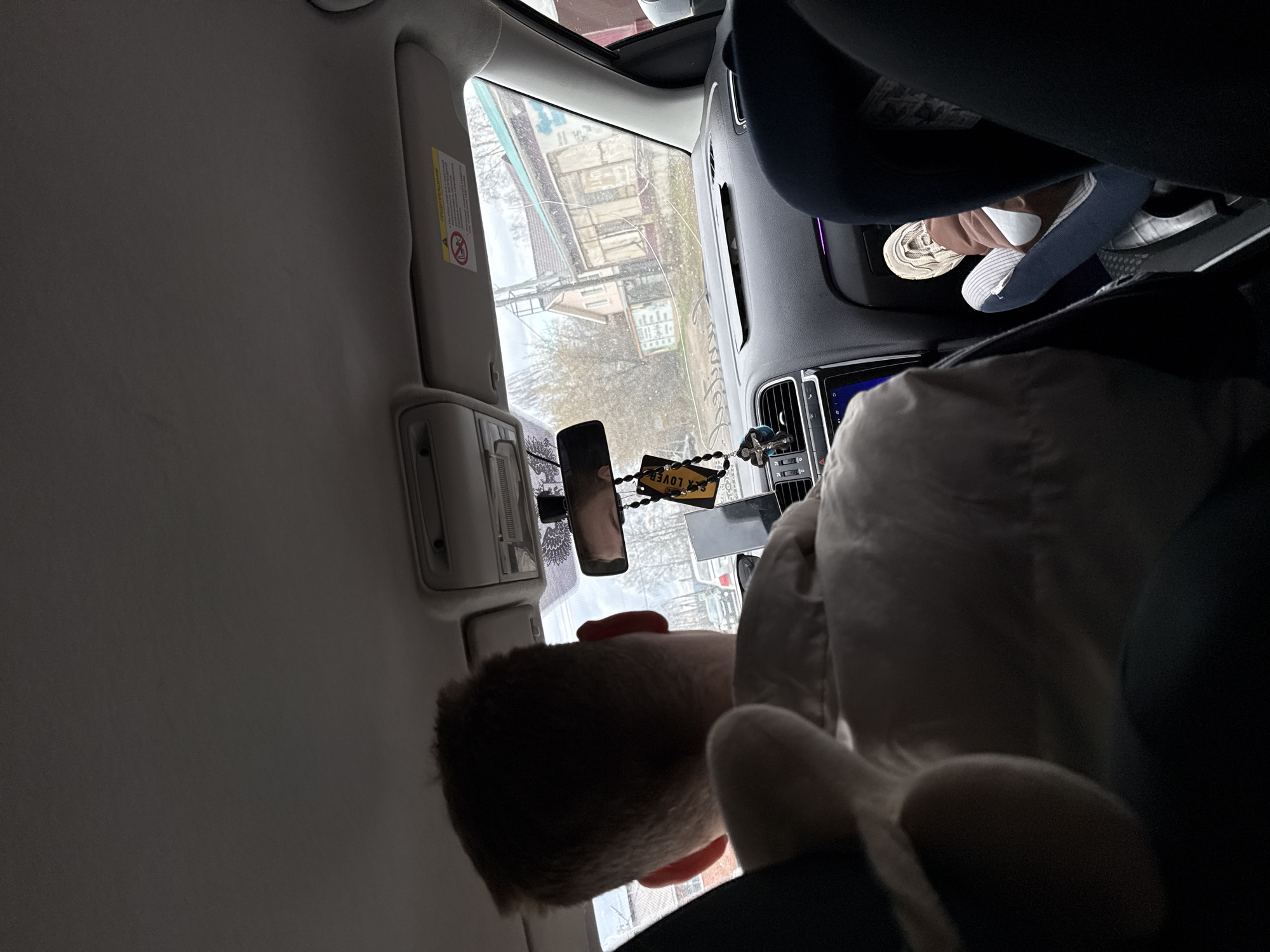 The photograph, taken from the interior of a car, shows a boy looking out the window. Outside, buildings and trees suggest an urban environment, consistent with Chaikovsky, Russia. A rear-view mirror and car interior details, like the sun visor, are visible. Another individual, with sock-clad feet, is seated in the backseat.