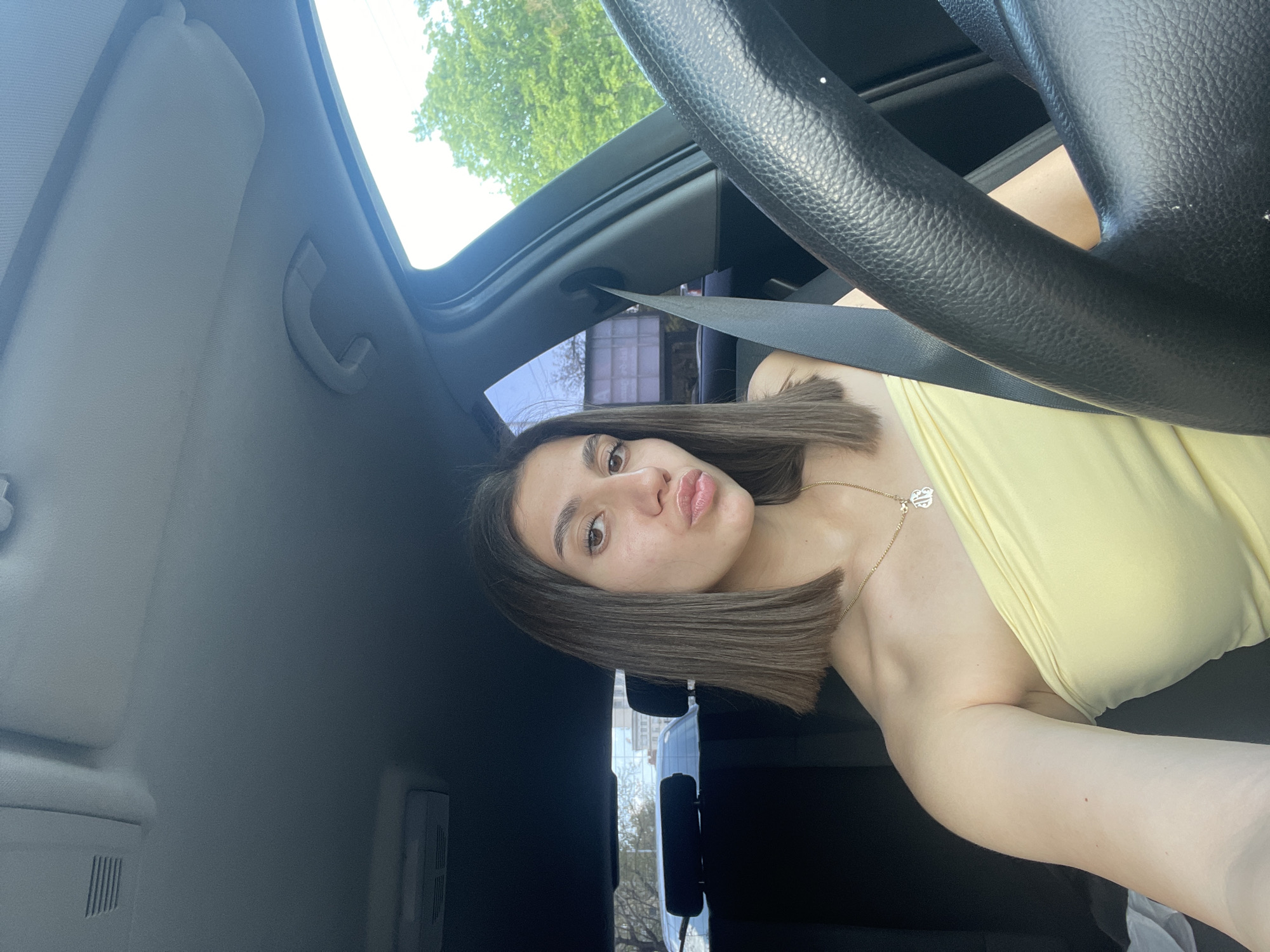 A young woman is featured in a selfie taken inside a car. She is positioned from the chest up, looking directly at the camera with a playful expression, pouting her lips slightly. She has straight, shoulder-length, dark brown hair and wears a light yellow strapless or off-the-shoulder top. A delicate gold chain necklace with a small heart-shaped pendant adorns her neck.
The setting is the interior of a vehicle, appearing to be from the passenger side, though a driver's perspective in a right-hand drive car is also possible. A black, textured steering wheel is partially visible on the right side of the frame, along with a seatbelt crossing her chest. Above, parts of the car's light-colored headliner, a grab handle, and a sun visor are visible. Through what appears to be a sunroof or moonroof, and the car's side windows, bright sunlight illuminates the scene, revealing green tree leaves and a clear sky, indicative of a sunny day. In the background, beyond the car windows, blurred architectural structures and more trees suggest an urban or suburban outdoor environment.
The activity is posing for a photo. The time of day is clearly daytime, sunny, and bright. The overall emotion conveyed by the woman is confident and playful. No distinct text is visible on her person or within the car's interior. The location context for this image is Sofia, Bulgaria.