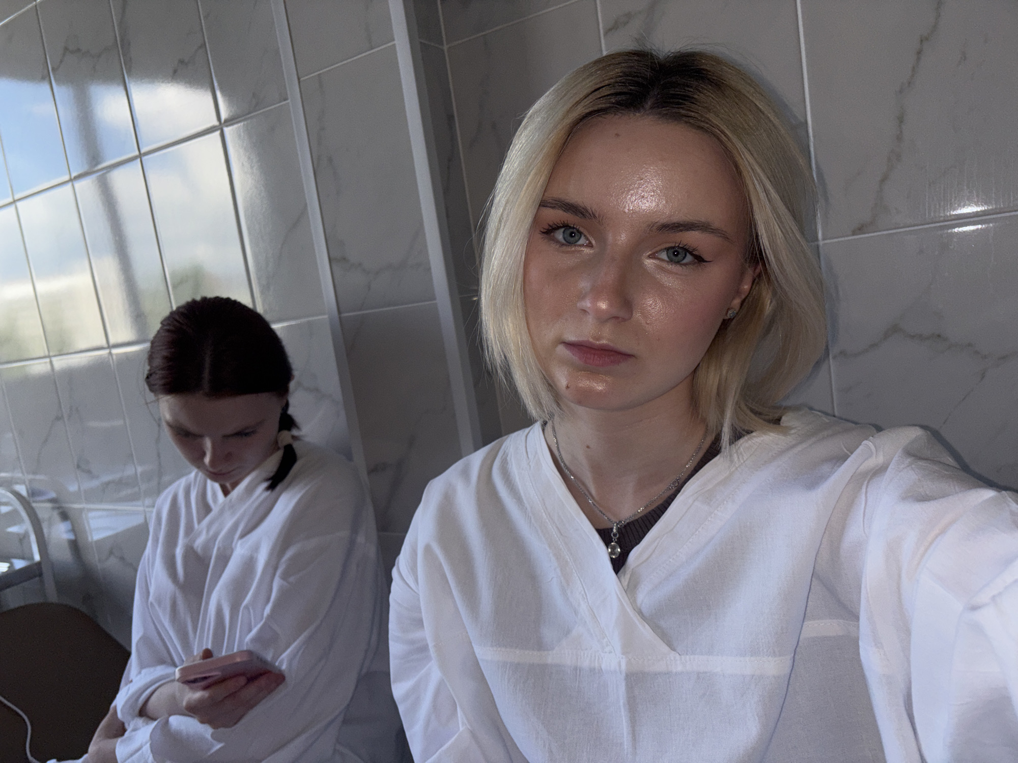 The image captures two young women indoors, likely within a medical or clinical environment such as a hospital room or a clean, tiled space. In the foreground, a blonde woman with shoulder-length hair and light blue eyes is taking a selfie, looking directly at the camera with a neutral expression. She is wearing a white V-neck top, which resembles a lab coat or patient gown, and a silver necklace with a small pendant. Her skin appears naturally luminous.
To her left and slightly behind her, another woman with dark hair, tied back, is visible. She is also dressed in a white garment, possibly a patient robe, and is focused on a pink smartphone she holds in her hands, with her face partially obscured as she looks downwards.
The setting features light-colored, possibly white or light gray, tiled walls, some displaying a subtle marble pattern. Reflections of a bright sky with clouds are visible on the tiles, indicating natural daylight from a nearby window. A metallic structure, possibly part of a hospital bed or other medical equipment, can be seen in the left background. The overall attire and environment strongly suggest a healthcare facility. No text is visible in the image.