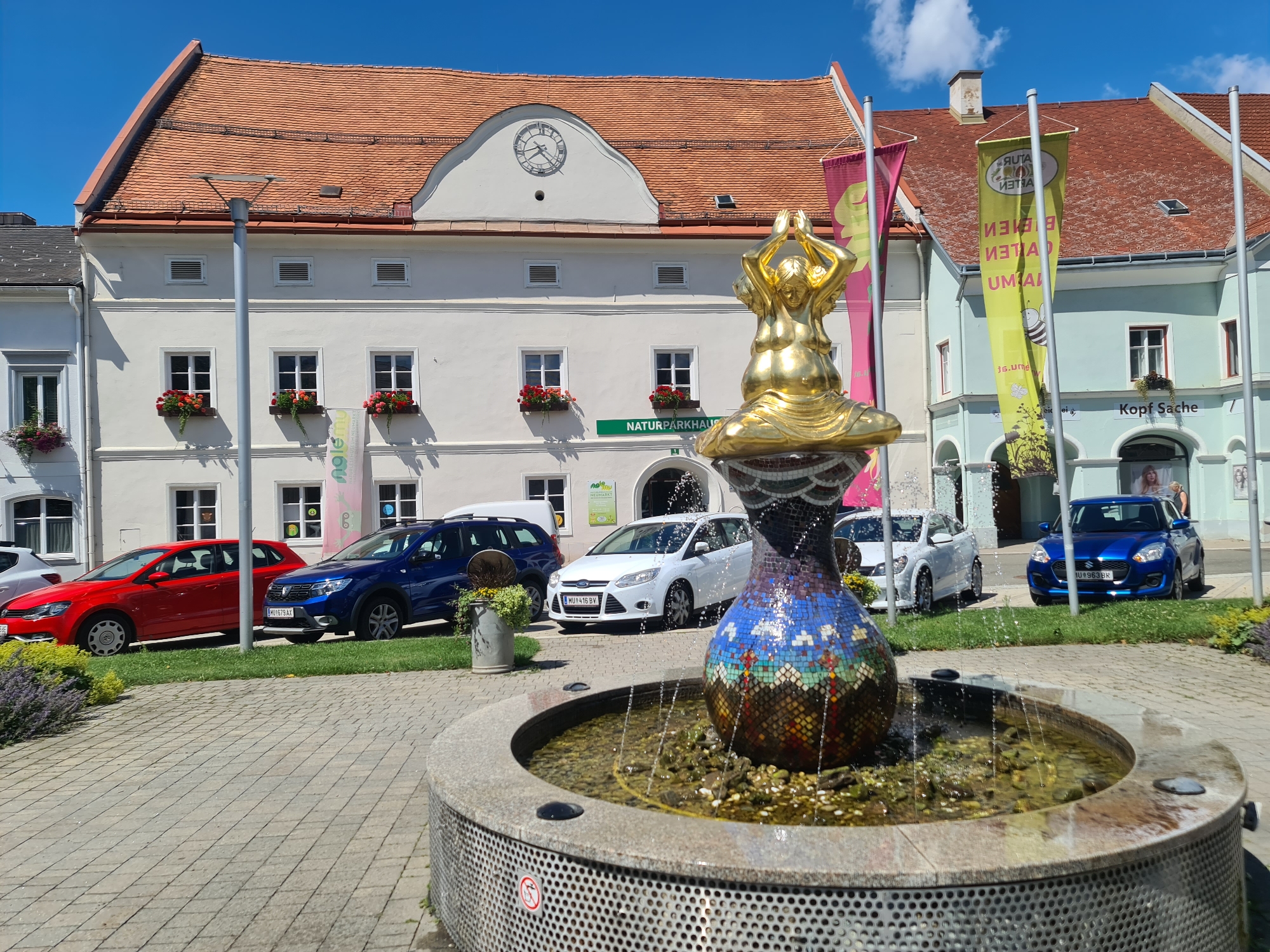 The image depicts a public square with a fountain in the foreground, located in Sankt Marein bei Neumarkt, Austria. The fountain is decorated with mosaic tiles and has water spraying upwards, with a golden statue of a seated figure on top. Behind the fountain, a street runs parallel to a row of buildings. Several cars are parked along the street, including a red sedan, a blue SUV, a white hatchback, and a blue compact car. Two flags are visible on poles to the right of the fountain, one pink and one yellow with text and graphics. The buildings are multi-story, with tiled roofs and visible windows. One building has a clock face on its facade. In the background, there is a clear blue sky with some white clouds. The overall scene suggests a sunny day in a town center.