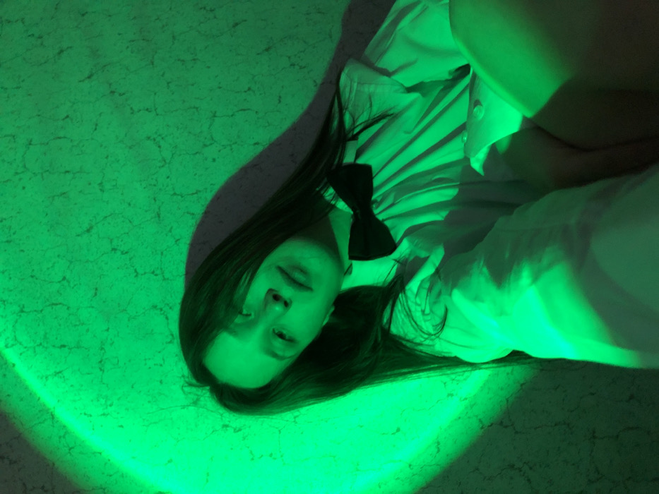 A woman lies on a cracked green surface. She wears a white shirt with a black bow tie. Her eyes are closed, and her head is tilted. The surface is illuminated by a curved green light. Her hair is long and dark, spread out on the surface. Her arm is bent and rests on her chest. The overall tone is eerie and dreamlike.