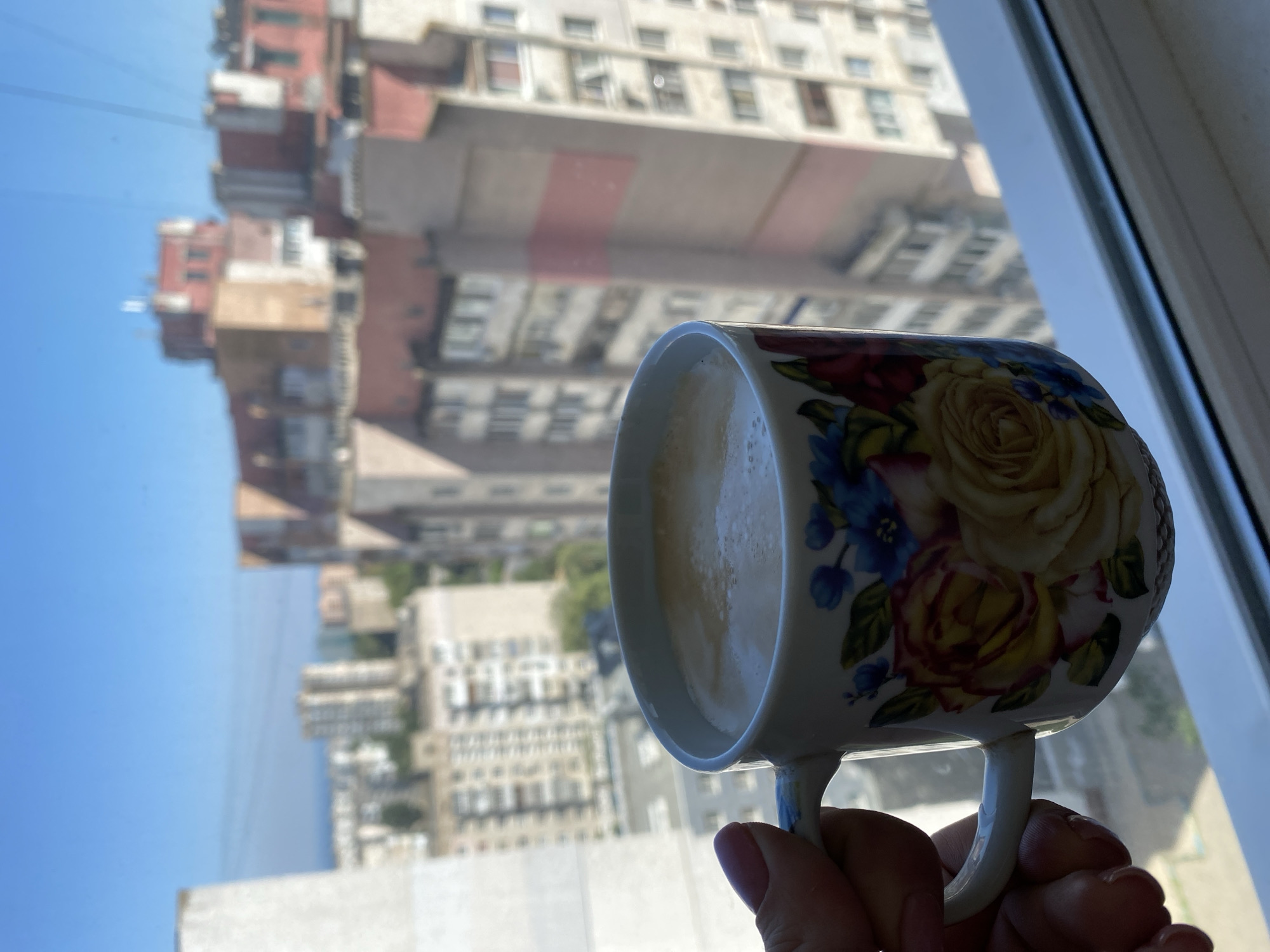 A hand with a pink fingernail holds a white mug decorated with colorful flowers, including yellow roses and blue blossoms, filled with a light brown beverage, likely coffee, with foam on top. The mug is positioned in the foreground, obscuring the bottom portion of a window frame. Through the window, a cityscape is visible under a clear blue sky. The background features several multi-story apartment buildings, some with beige and red facades, and one with pink accents. The architecture suggests an urban environment, consistent with Chornomorsk, Ukraine. The lighting indicates it is daytime, with the sun illuminating the buildings. The overall scene captures a moment of enjoying a drink while looking out at the city.