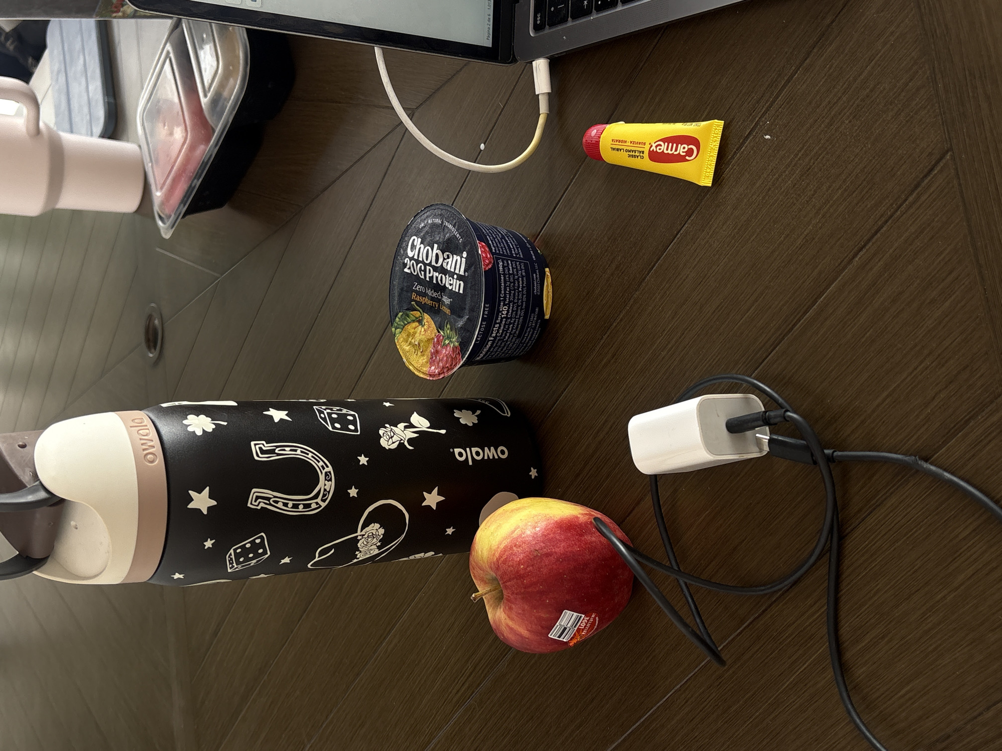 A top-down shot on a wooden floor reveals several items. A laptop occupies the top edge of the frame, with a white charging cable plugged into it. A container of Chobani 20G Protein yogurt and a tube of Carmex lip balm are near the laptop. An Owala water bottle, with a black body decorated with white icons, rests next to an apple. A white charging block with black cables sits close to the apple. The scene appears to be indoors. There is no indication of the location being in Puebla, Mexico, or any people present.
