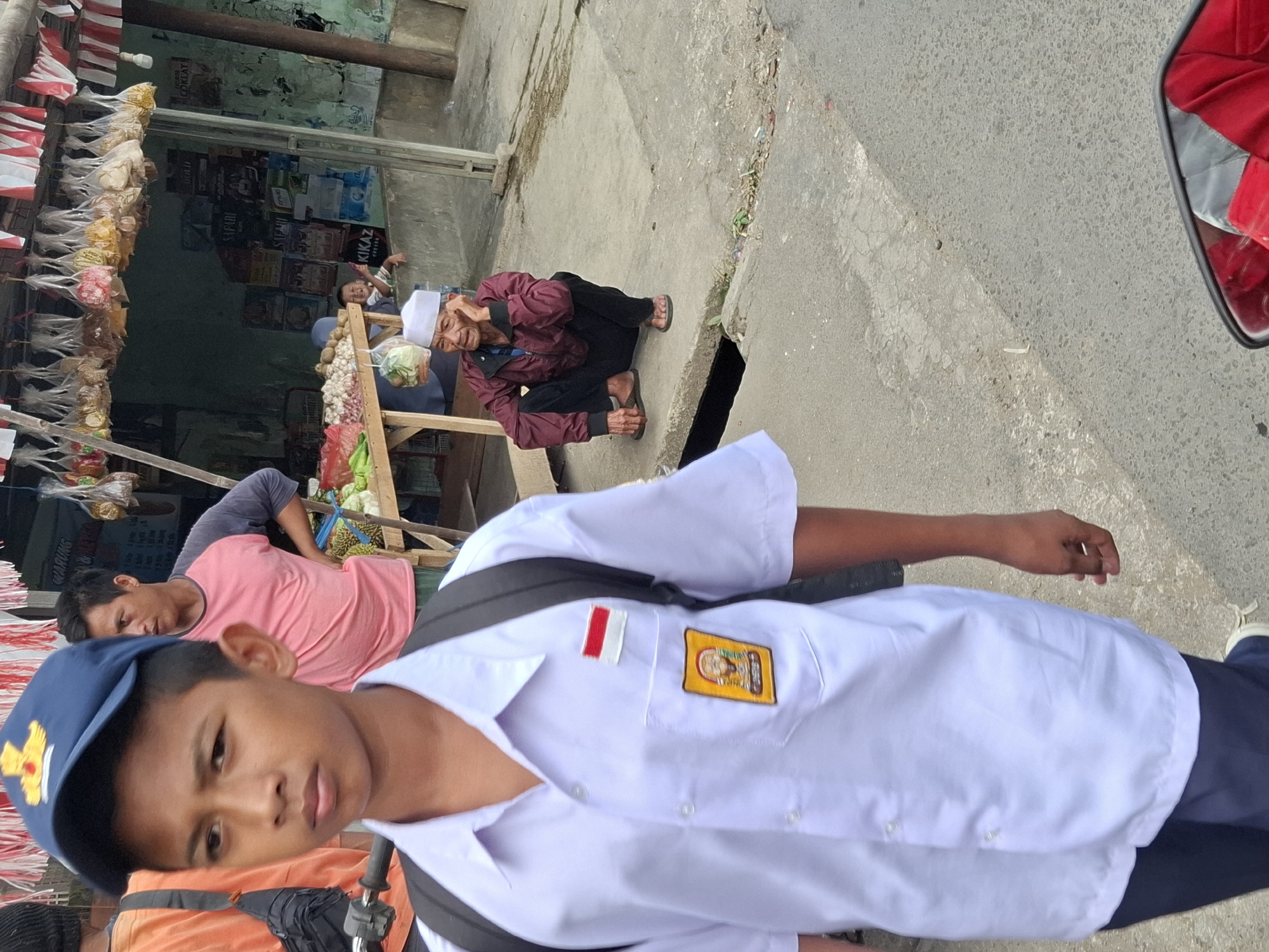 In Balekambang, Indonesia, a young boy wearing a blue cap and a white school uniform with a backpack walks forward, his face visible and looking towards the camera. To his right, a man in a pink shirt stands with his hand on his hip, looking away. Further back, an elderly man in a maroon jacket and white cap squats on the concrete, resting his hand on his face. Behind the elderly man, a small child peeks out from behind a market stall adorned with red and white flags. The market stall displays various packaged goods and fresh produce. The scene unfolds on a paved street with a gutter, under a partially covered market area.