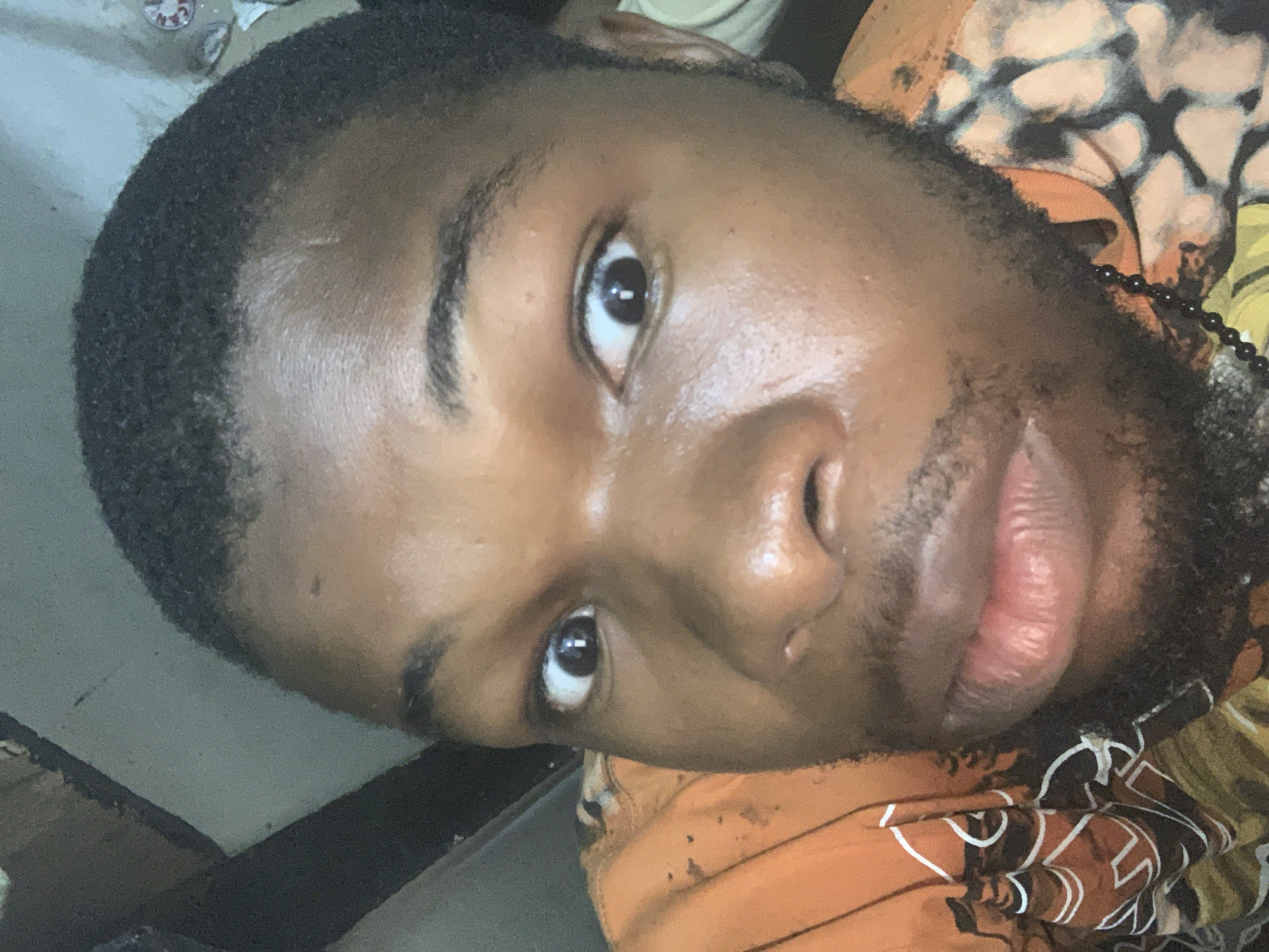 A close-up, medium-shot photograph depicts a Black man's face, angled slightly toward the viewer. He has short, dark hair, brown eyes, and a trimmed beard. His skin appears somewhat shiny. He is wearing a patterned orange and tan shirt. A beaded necklace is visible around his neck. The background is indistinct. The location, Fegge, Nigeria, is not directly discernible from the image.