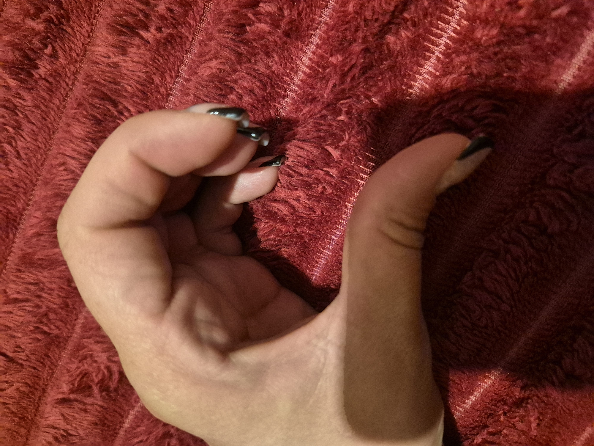 A close-up, medium shot shows a person's left hand, with the fingers curled to form a heart shape. The hand is positioned on a textured, dark red surface, possibly a blanket or rug. The person's nails are painted black. The background is out of focus, with the red texture filling most of the frame. There is no discernible urban environment or location information in the image.