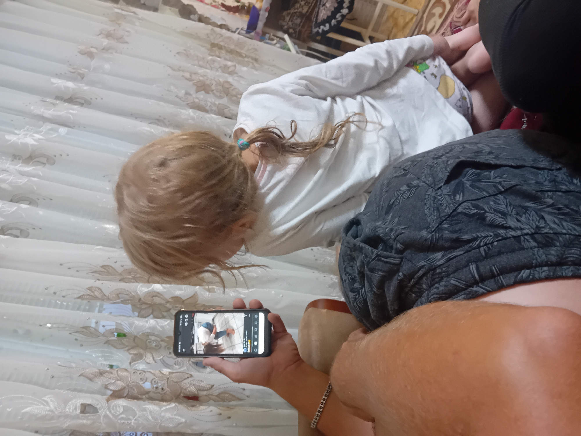 The media file shows a close-up of a young girl with blonde hair, wearing a white long-sleeved shirt and patterned shorts. She is looking down at a smartphone held by an adult whose arm and shoulder are visible in the foreground. The adult is wearing a dark patterned shirt and a silver bracelet. The smartphone displays a video or image of another person, possibly the girl, in a similar pose.
The scene appears to be indoors, with a white curtain with floral embroidery visible in the background. The lighting suggests it could be daytime, but no specific time is indicated. The overall mood seems relaxed and domestic, with the focus on shared screen time. The girl's hair is tied back with a small blue and pink hair tie, and a few strands are loose around her face. The adult's skin tone is light, and their arm is slightly tanned. The phone is a modern smartphone with a visible screen interface.
There is no visible text in the image itself, apart from what might be on the smartphone screen, which is too small and blurry to read. The image is oriented sideways. The setting is consistent with a home environment in Chuhuiv, Ukraine.