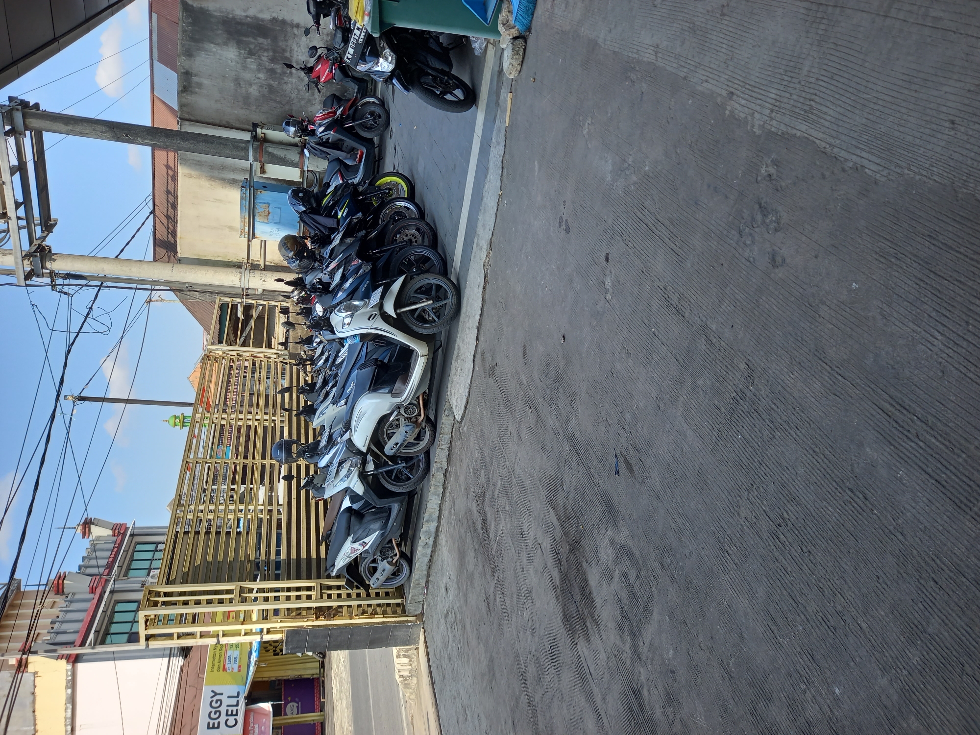 The image shows a row of parked motorcycles in front of a building in Samarinda, Indonesia. The motorcycles are lined up on a concrete surface, with a yellow metal fence behind them. The building appears to be a commercial establishment, with signage visible that reads "EGGY CELL". The sky is blue with some clouds. The overall scene depicts a typical street view in a city environment.
