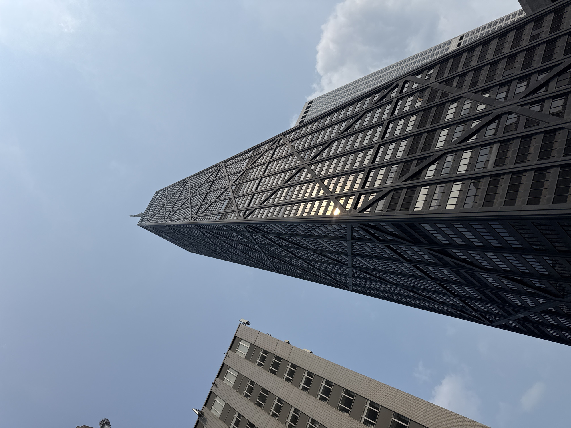 The image shows a low-angle view of the John Hancock Center in Chicago, Illinois, United States of America. The skyscraper is characterized by its distinctive X-braced exoskeleton and dark glass facade, which reflects the bright blue sky and some scattered clouds. To the left of the John Hancock Center is another tall building with a lighter-colored facade and a grid of windows. The perspective emphasizes the height and scale of the skyscrapers against the sky.