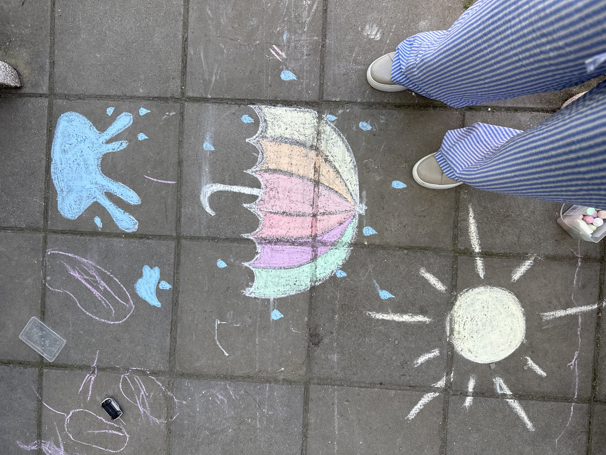 The image shows a grey tiled ground with chalk drawings. There is a blue splattered handprint, a multi-colored umbrella with blue raindrops, a white sun, and some other indistinct pink and blue drawings. A pair of legs wearing blue and white striped pants and beige shoes are partially visible. A clear plastic container with colored candies is positioned near the shoes. A small black object, possibly a toy car, and a clear container are also on the ground. The location appears to be an outdoor paved area, possibly in a city.
