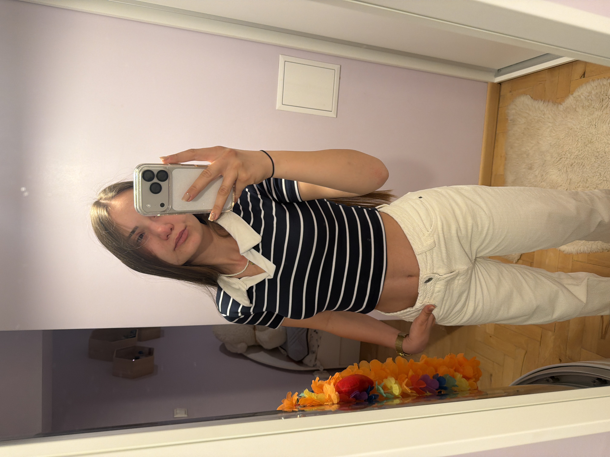 A young woman is taking a selfie in a mirror. She is wearing a striped crop top, light-colored pants, and a bracelet. In the reflection, there is a room with purple walls, a white square decoration, a white rug, and a garland. The location is not discernible as Buhovo, Bulgaria.