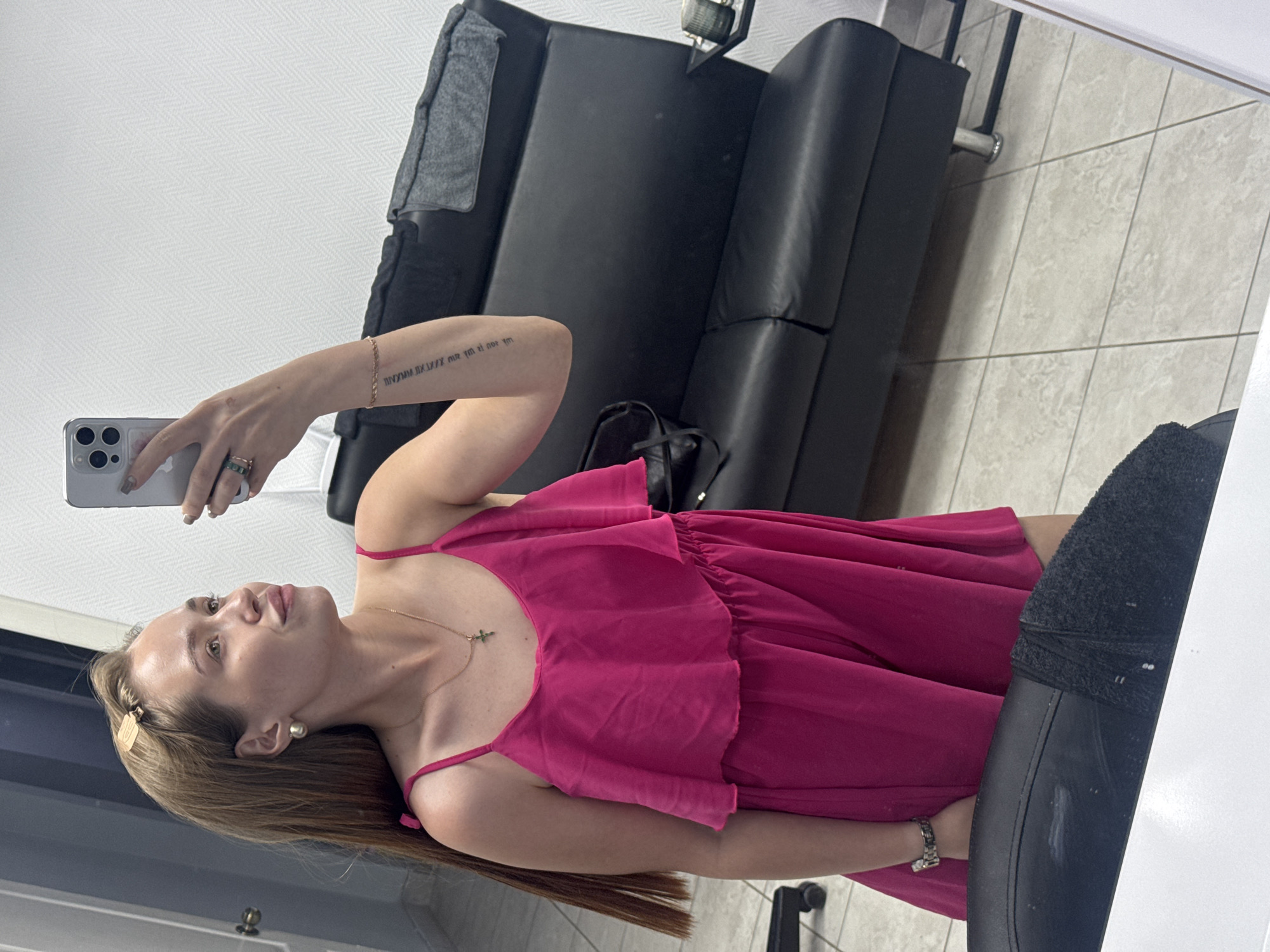 A woman is taking a selfie in a room. She is wearing a pink dress and jewelry. She is holding a phone in her left hand, and the phone case is gray. The room has a black leather couch and is reflecting in a mirror. It's possible the location is Chaikovsky, Russia.