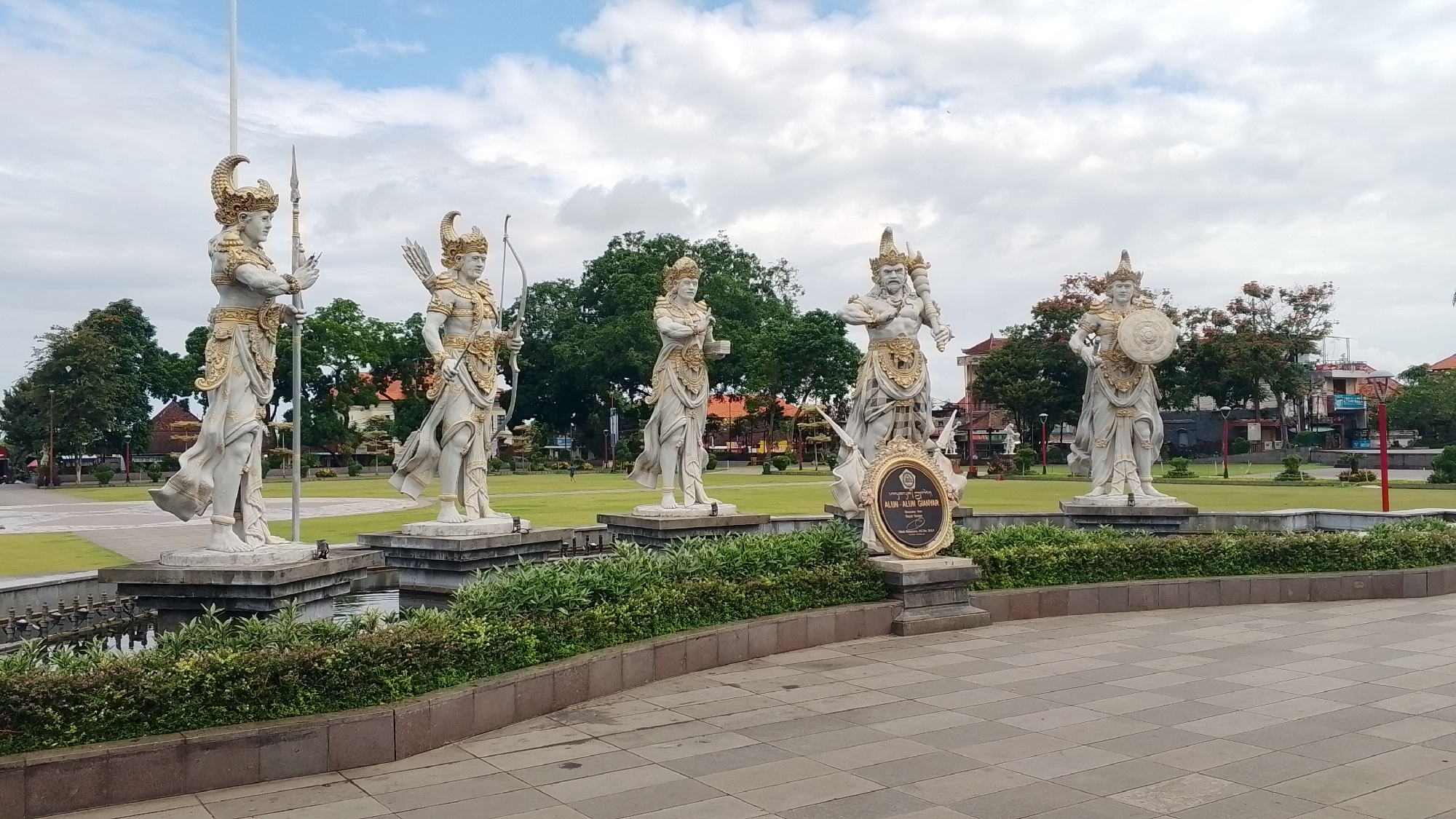 Five large, white stone statues of figures in ornate golden attire stand in a row on stone pedestals. The statues are of warriors, each holding a different weapon or shield; the leftmost statue holds a spear, the second from the left holds a bow and arrow, the middle statue holds a goblet, the second from the right holds a mace, and the rightmost statue holds a shield. The statues are located in an outdoor plaza with lush green grass and trees in the background. A small body of water with decorative bushes is in front of the statues, and a tiled pathway is in the foreground. The sky is cloudy with patches of blue. A framed plaque with writing in an unknown language is situated in front of the middle statue.