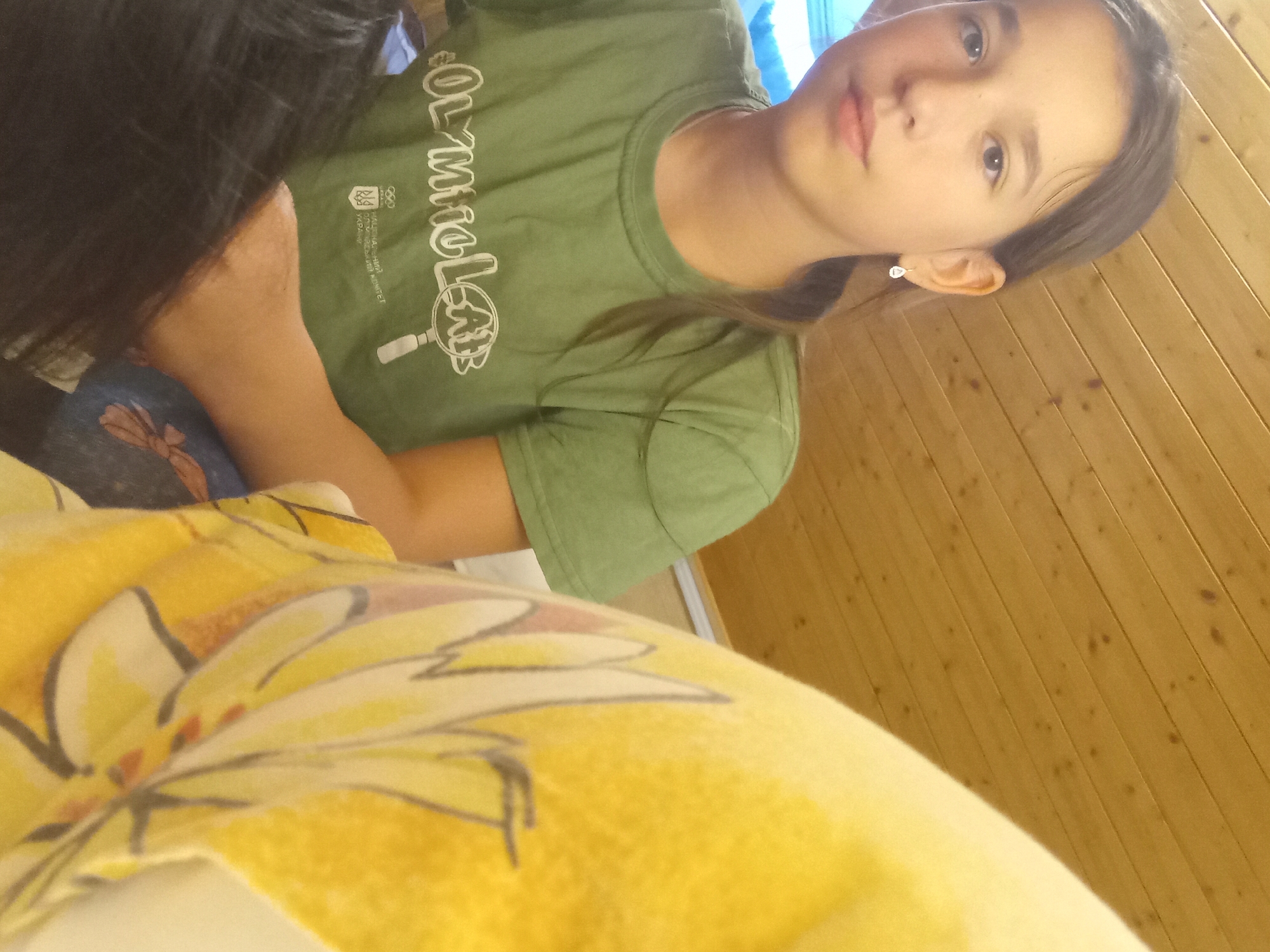 A young girl is pictured in a close-up, selfie-style photograph. She has light brown hair pulled back in a ponytail and is wearing a green t-shirt with white lettering and a Ukrainian Olympic committee logo. She is also wearing a small, silver, tear-drop shaped earring in her left ear. The background consists of a wooden, sloped ceiling. The lower right portion of the image is occupied by a yellow fabric with a floral pattern. The image appears to have been taken indoors in a room with wooden paneling on the ceiling, which is common in some Slovakian homes, particularly in areas like Zuberec, a village known for its traditional wooden architecture.