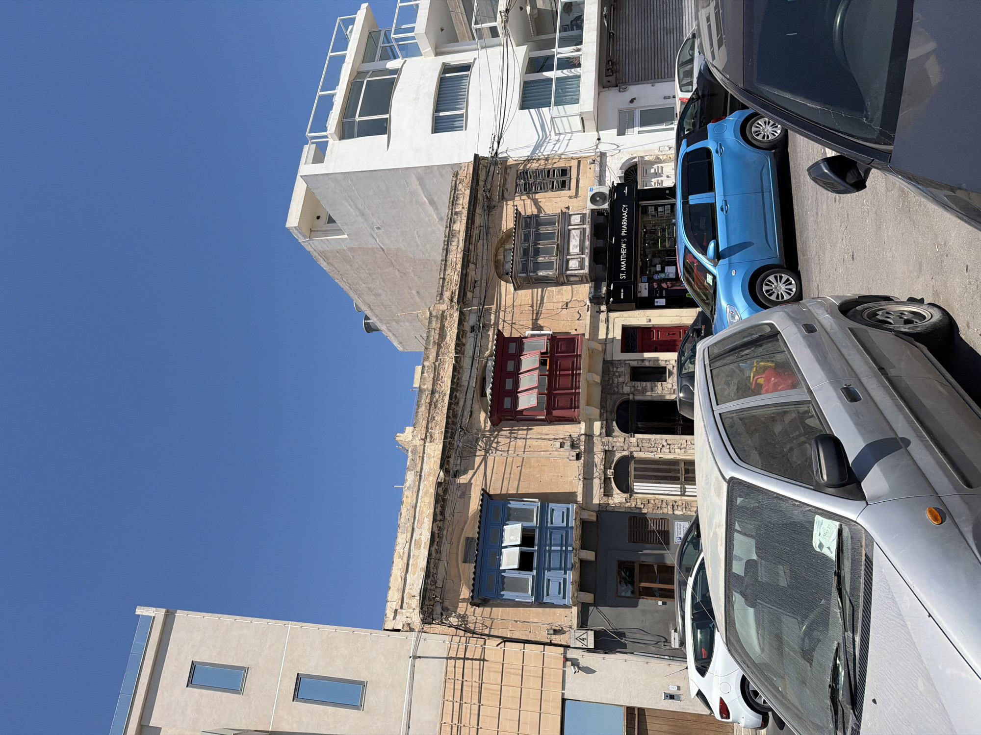 This image captures a street scene in Gżira, Malta, under a clear blue sky. The foreground is dominated by the front of a dusty silver car, its windshield bearing a parking ticket. To its right, the dark roof and side mirror of another car are visible. Behind these cars, a bright blue compact car is parked on the street.
Further back, a row of buildings forms the backdrop. The central building is an older, two-story structure with a weathered, pale yellow facade. It features two distinct enclosed balconies on the upper floor: one painted a vibrant blue with open windows, and another to its right painted red, also with open windows. Below these balconies, the ground floor of this building has several arched doorways and shop fronts. One prominent shop is labeled "ST. MATTHEW'S PHARMAÇY" with a black awning.
To the left of the central older building is a taller, modern, light-colored building with rectangular windows. To the right of the older building, another modern white building rises, featuring multiple levels with balconies and large windows, some with curved edges. Overhead, numerous electrical wires crisscross the scene, connecting the buildings. The overall impression is a typical urban street in Malta, showcasing a mix of architectural styles and parked vehicles.
