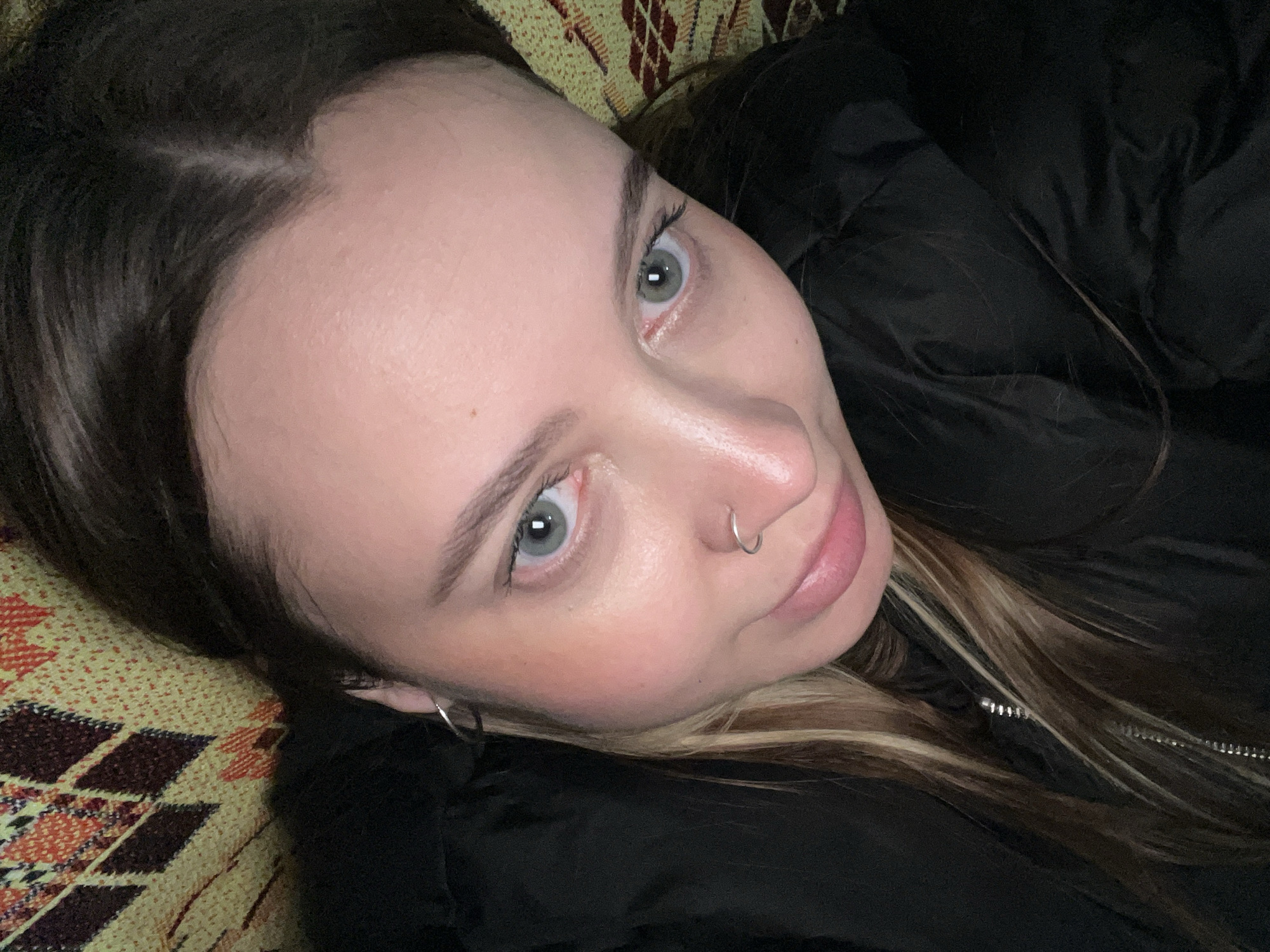 A close-up selfie of a woman with light skin and grey eyes. She has dark brown hair with some blonde highlights and a silver nose ring. She's wearing a black puffy jacket and lying on a patterned surface. There is no information in the image to determine a specific location or connection to Novopokrovskaya, Russia.
