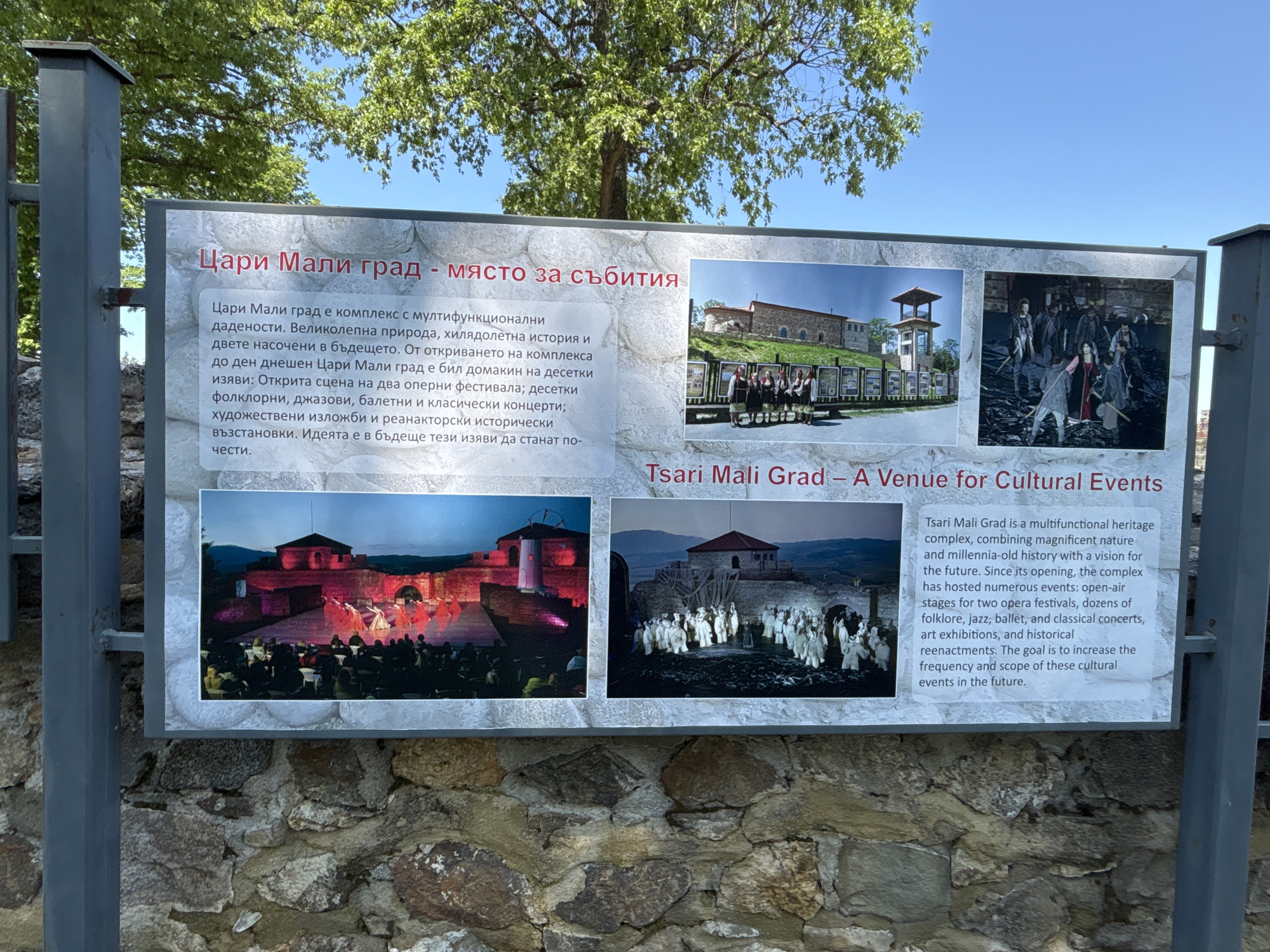 A rectangular information sign, secured by gray metal posts, is positioned outdoors against a stone wall background in Belchinski Bani, Bulgaria. The sign's surface displays a white textured background, mimicking stone, bordered by a darker gray frame.
The sign is titled in red capital letters in both Bulgarian and English. The Bulgarian title, "Цари Мали град - място за събития," appears at the top left, translating to "Tsari Mali Grad - a venue for events." Below this, a detailed description in Bulgarian text outlines Tsari Mali Grad as a multifunctional heritage complex, highlighting its natural beauty, ancient history, and future vision. It mentions the complex has hosted numerous events, including opera festivals, folklore, jazz, ballet, classical concerts, art exhibitions, and historical reenactments, with plans to expand these offerings. The English translation of the title, "Tsari Mali Grad – A Venue for Cultural Events," is visible towards the bottom right of the sign. A corresponding English text block, located on the right side of the sign, reiterates the information provided in the Bulgarian text.
Four photographic images are arranged on the sign: two horizontally aligned at the top and two horizontally aligned at the bottom.
* The top-left image features a group of individuals in traditional Bulgarian attire standing in front of a stone building with a prominent bell tower under a clear blue sky. Several display boards are visible in the background.
* The top-right image depicts a historical reenactment or performance with a group of people in period costumes, some carrying props resembling weapons, on a dark, textured stage.
* The bottom-left image shows an evening outdoor stage performance with dancers in vibrant costumes, illuminated by red and pink stage lighting, performing for an audience seated in chairs. The stone architecture of the complex forms the backdrop.
* The bottom-right image presents another outdoor performance or reenactment, with a group of individuals dressed in white costumes on a stage. The stone complex is visible in the background under what appears to be ambient blue lighting.
The metal posts supporting the sign are a uniform gray. Behind the sign, a section of a rough-hewn stone wall is visible. Above the sign, the bright blue sky with scattered white clouds is prominent, accompanied by the lush green foliage of a deciduous tree, indicating a daytime outdoor setting.