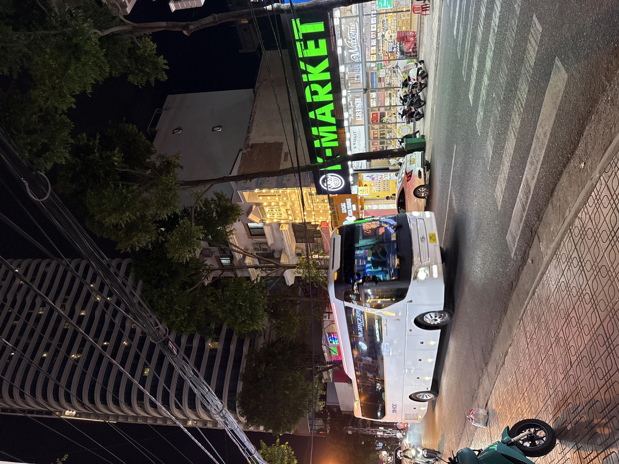 The image shows a street scene in Nha Trang, Vietnam at night. A large white bus with its headlights on is driving down the street. To the right of the bus, there is a K-MARKET convenience store with bright green lettering on its sign. The store appears to be open, with people inside and goods displayed on shelves. Several motorcycles are parked in front of the store. In the background, there is a tall, modern building with horizontal stripes and lit windows. Trees line the street, and there are streetlights and overhead wires. The sidewalk is made of patterned tiles, and a green electric scooter is parked on the sidewalk in the foreground. The street has a pedestrian crossing marked with white stripes.