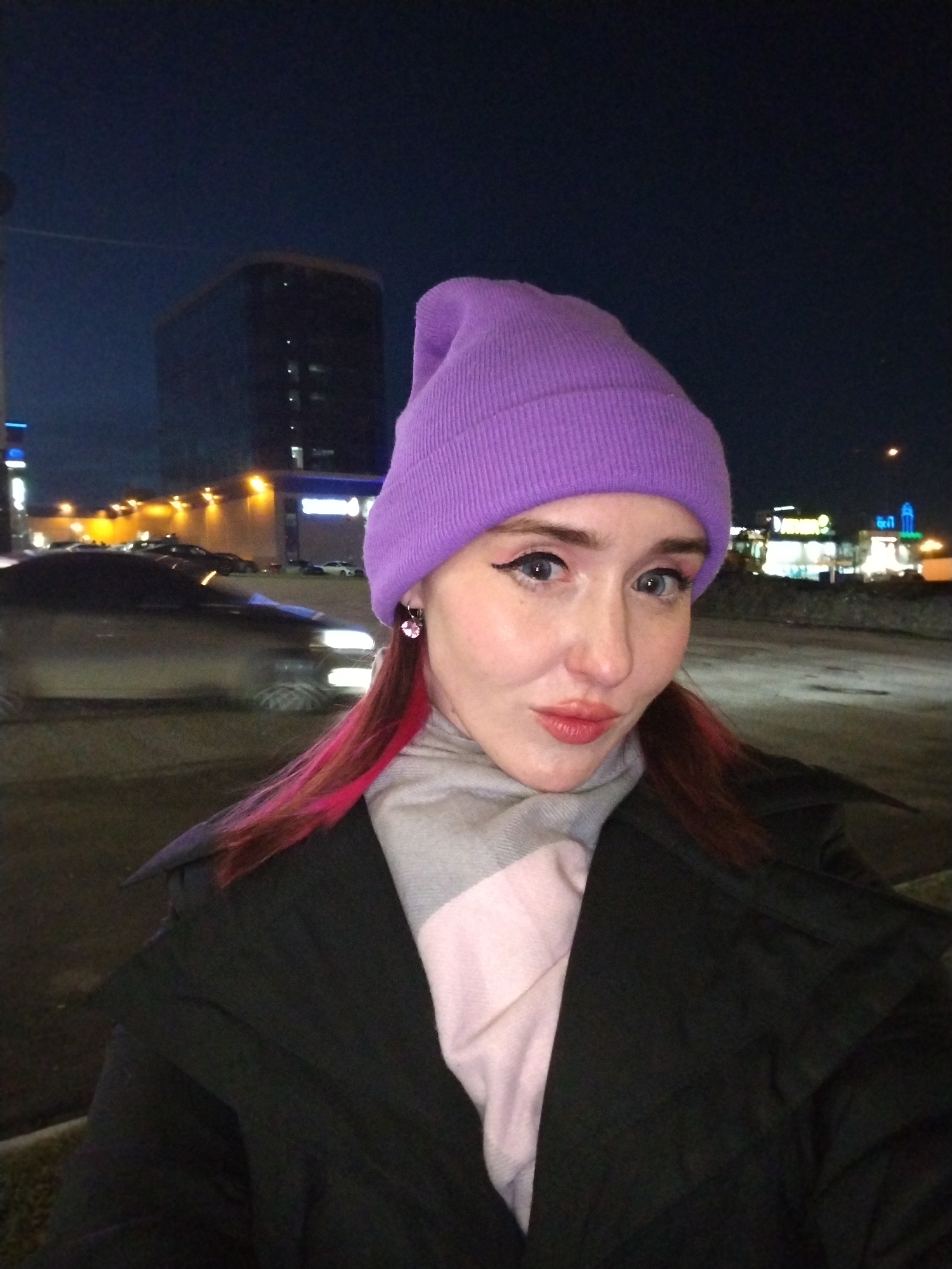 A woman is standing outside at night. She is wearing a purple beanie, a grey and pink scarf, and a black coat. She has dyed pink streaks in her hair and is wearing pink earrings. She is looking at the camera. In the background, there are lights and buildings. The location is Izhevsk, Russia. The buildings visible in the background include the Mercure Izhevsk hotel on the left and the Arbat hotel to the right. A blurry car with headlights on is moving in the background.