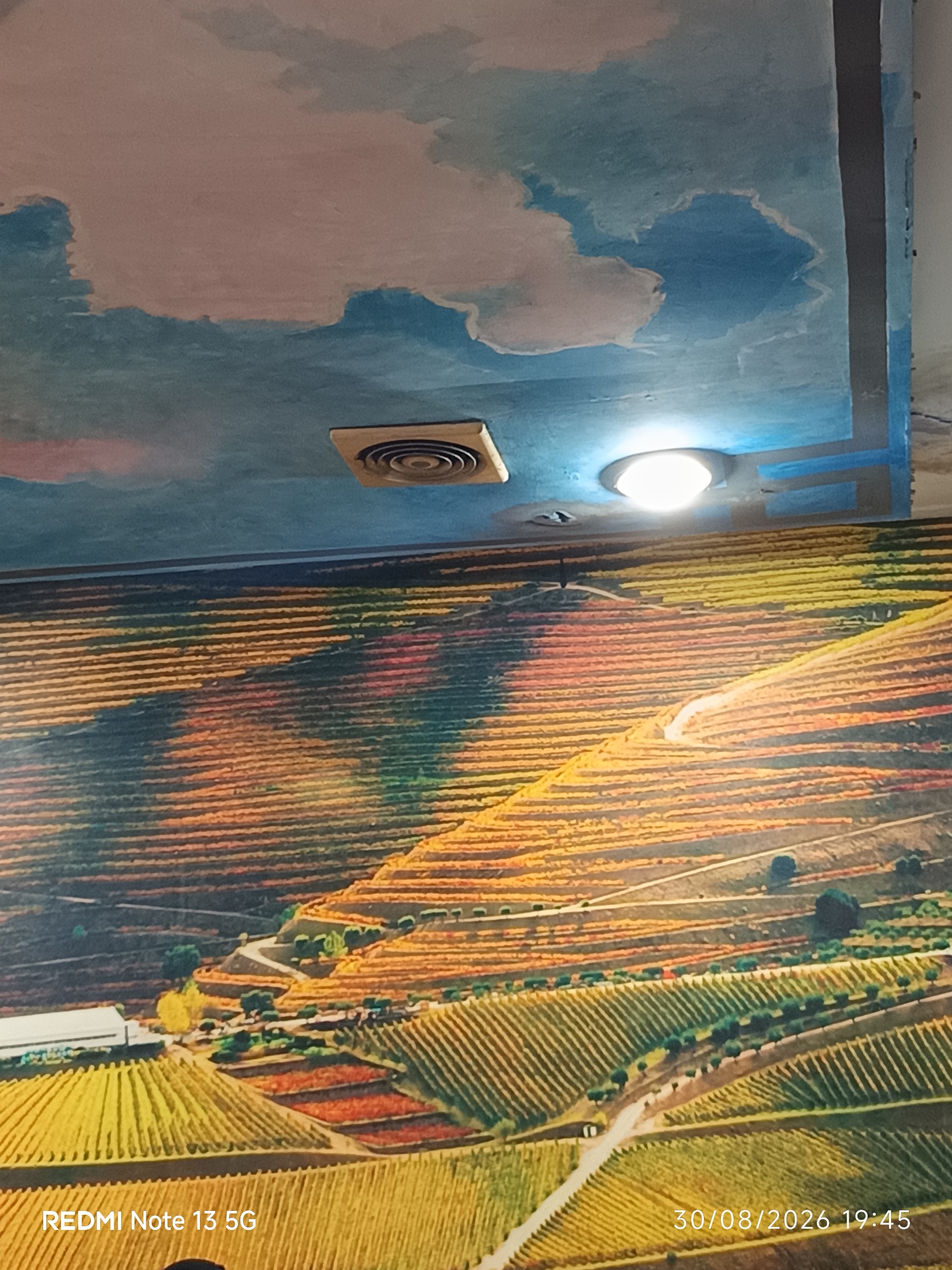 The image displays a mural or painting on a ceiling and wall, depicting a landscape of terraced vineyards. The upper portion shows a sky with clouds, rendered in shades of blue and pink, with a ventilation grate and a circular light fixture visible. The lower portion, on the wall, features a vibrant, stylized representation of rolling hills covered in vineyards. The rows of vines are depicted with alternating colors of yellow, orange, and red, suggesting different seasons or types of crops. A winding road snakes through the vineyard, and small clusters of trees and buildings are scattered throughout the landscape. In the bottom left corner, the text "REDMI Note 13 5G" is present, and in the bottom right corner, the date "30/08/2026 19:45" is displayed. The location is stated as Kota Galuh, Indonesia, although this is likely a textual overlay and not visually represented within the artwork itself. There are no people or active interactions depicted in the image.