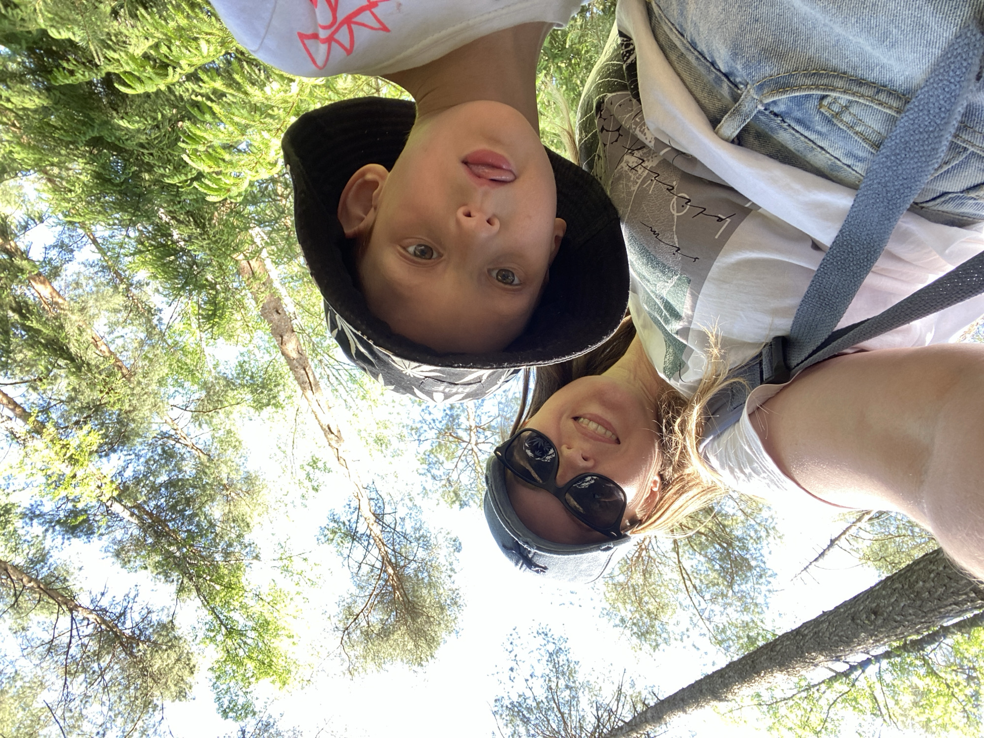 The photograph depicts two individuals from a low-angle perspective, appearing to be a selfie taken in an outdoor, forested environment, possibly in the city of Noviy, Russia. The first individual, presumably a child, is wearing a dark-colored bucket hat with a patterned design and a white t-shirt. The child is sticking their tongue out towards the camera. The second individual, likely an adult, is wearing sunglasses and a hat. The adult is smiling. The background consists of a canopy of trees with patches of sunlight filtering through. The photograph has a tilted orientation.