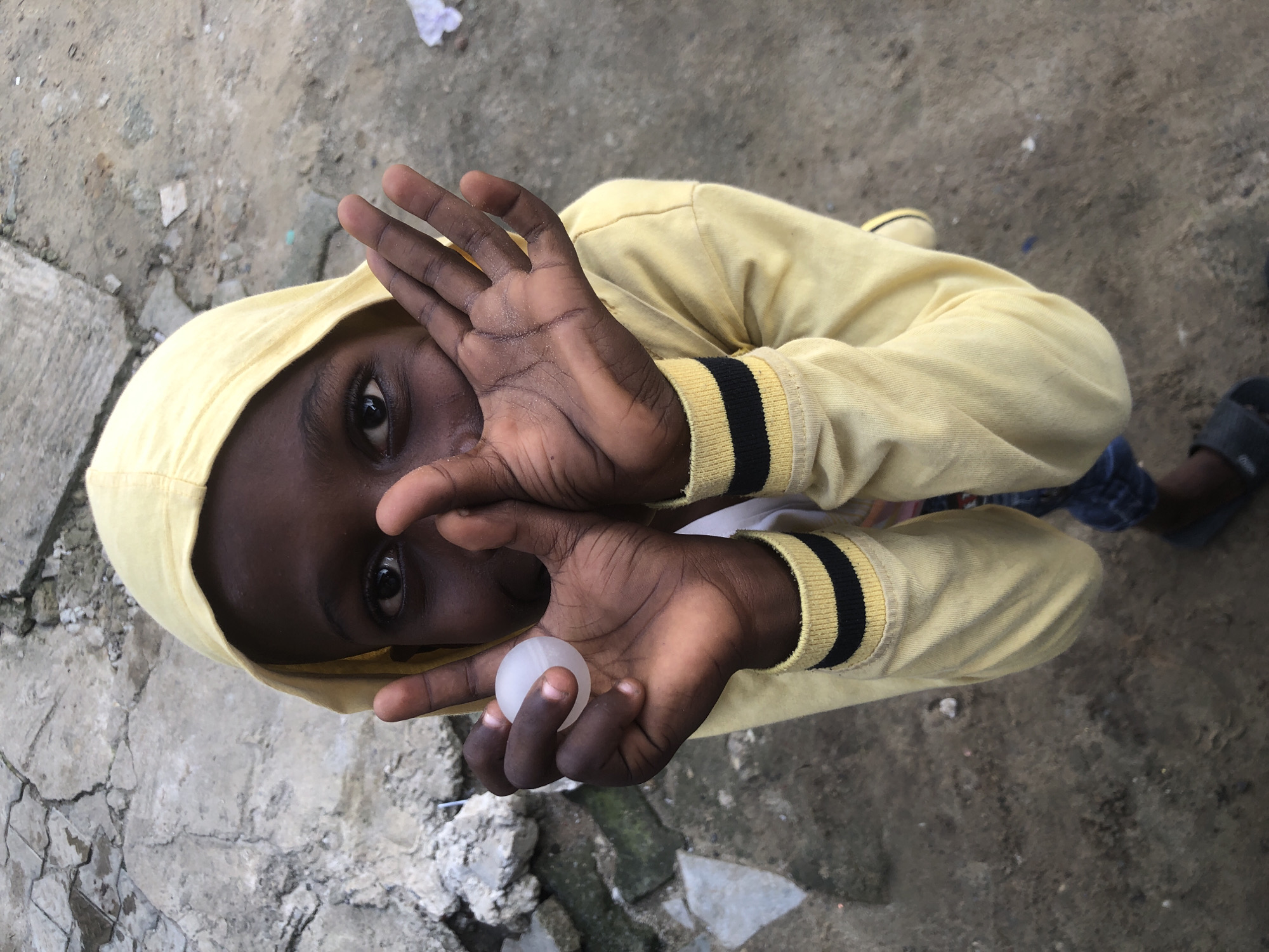 A high-angle medium shot shows a young Black child wearing a yellow hooded sweatshirt with black and yellow striped cuffs, standing outdoors on a dirt and gravel ground. The child is holding a small, translucent white sphere in their left hand, which is positioned in front of their face, partially obscuring their mouth and nose. Their right hand is raised and open, also in front of their face, with fingers spread. The child's eyes are visible, looking towards the camera. In the background, to the left, are large, rough grey stones and some weathered concrete or brick structures. The ground is a mix of dry dirt and small stones, with some debris visible. A small piece of white paper or fabric is on the ground to the right. The scene appears to be in Ado, Nigeria, characterized by its natural and somewhat unpaved environment.