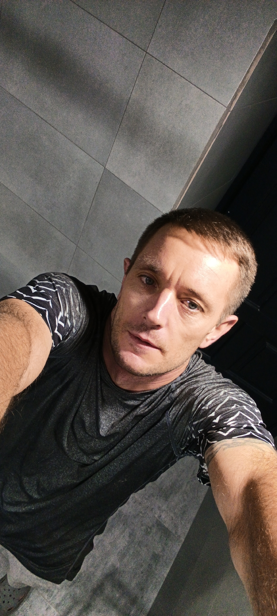 This is a medium-shot, high-angle, selfie-style photograph of a man indoors. The man is white, with short brown hair, blue eyes, and light stubble. He is wearing a dark gray t-shirt with a black and white patterned design on the sleeves. The background consists of a gray tiled wall and a dark ceiling. The lighting suggests an indoor environment, possibly a gym or a commercial space, with a strong light source casting shadows. The man is looking directly at the camera with a neutral expression. The photograph appears to be taken from a low perspective, looking up at the man. There are no other individuals or objects of note in the image. The location is not identifiable as Tashkent, Uzbekistan based on the visual information provided.