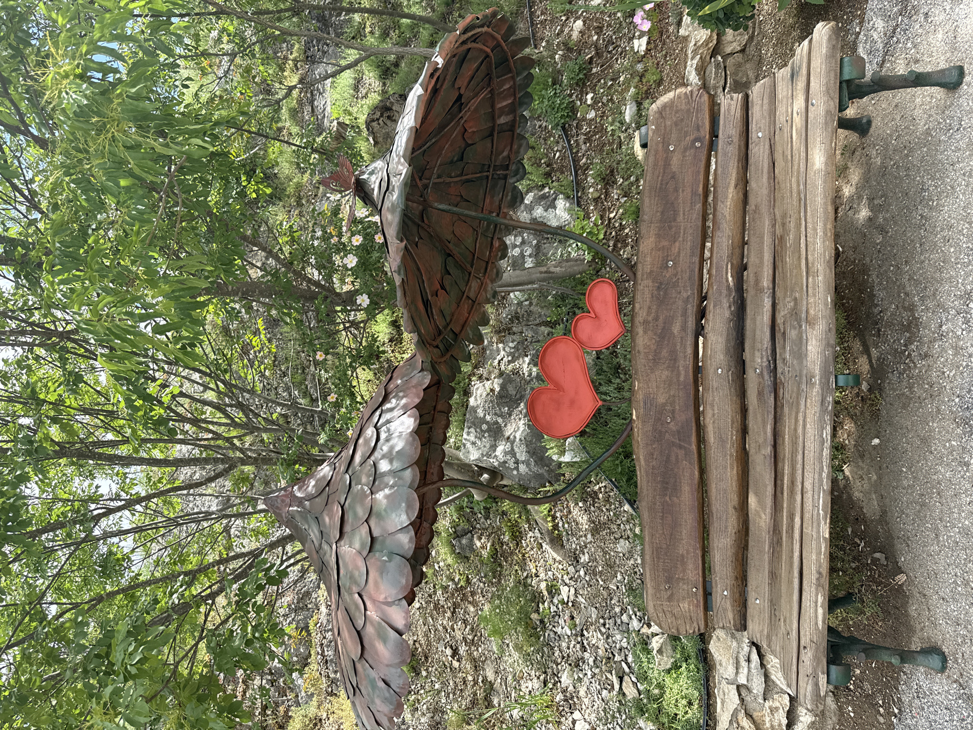 The image captures a scene in Lukovit, Bulgaria, featuring a rustic wooden bench situated outdoors amidst greenery and a gravelly surface. Adjacent to the bench is a metal sculpture resembling two peacocks, each crafted with a distinct fan-like tail. Two red heart-shaped decorations are positioned near the bench, adding a touch of sentimentality to the scene. The presence of lush trees and foliage in the background creates a natural and tranquil setting.