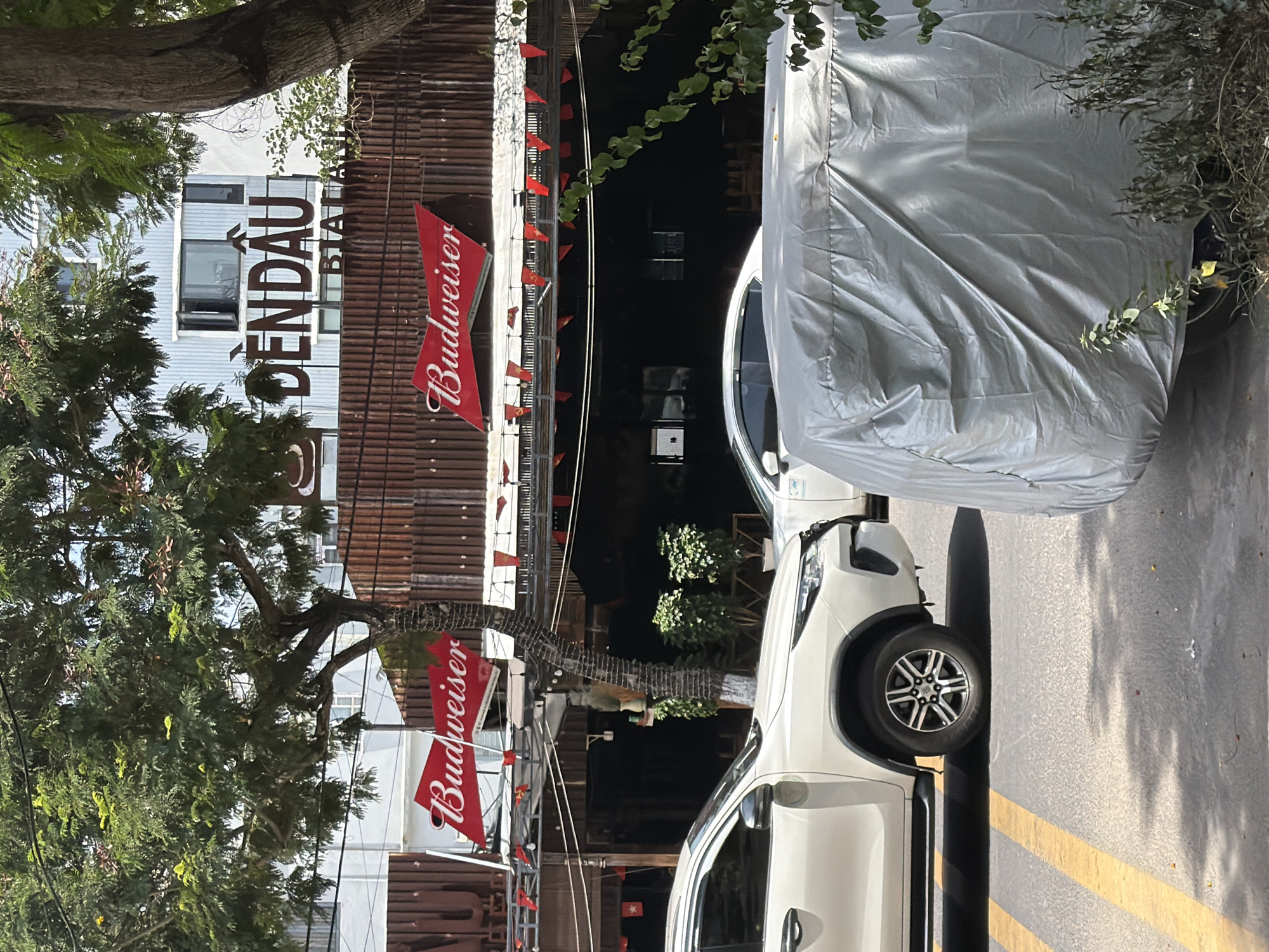 In the city of Đà Nẵng, Vietnam, this scene captures the exterior of a Budweiser-branded establishment. The building's facade features prominent red Budweiser logos and a sign with Vietnamese characters that read "DENDOAU BIA ANH." A white SUV is parked on the street in front of the establishment, partially obscured by a silver car cover. Trees with lush green foliage provide shade, casting dappled light onto the street and sidewalk. The environment suggests a commercial area with a focus on a beverage business.