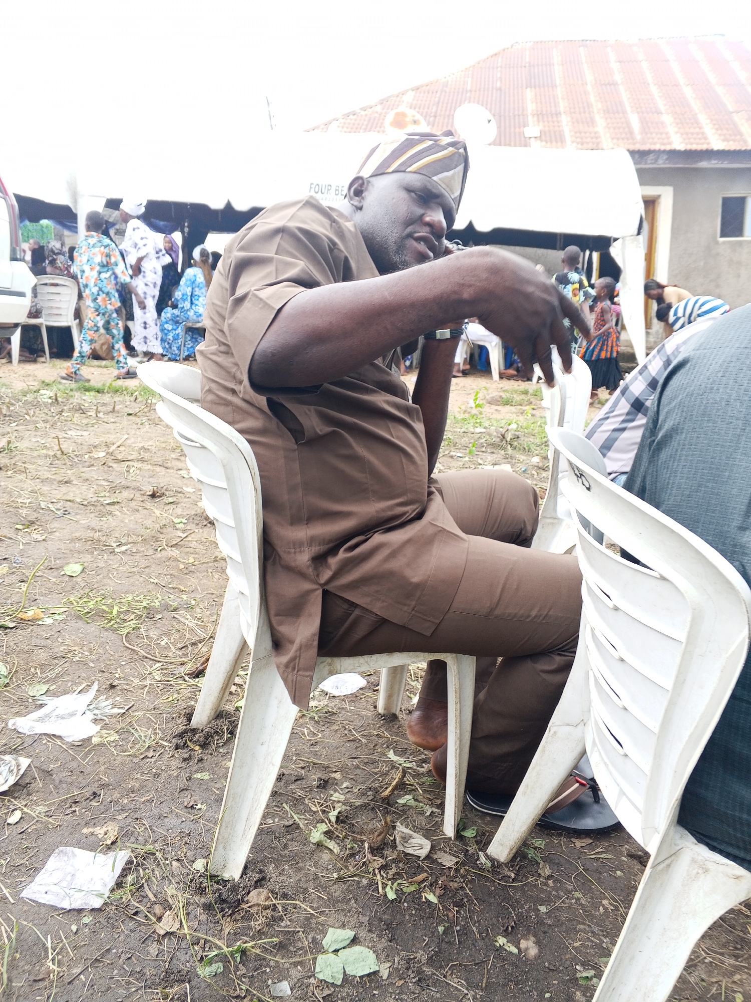 The image shows a man in traditional clothing sitting on a white plastic chair outdoors, with a tent and buildings in the background. He is wearing a brown outfit and a striped headwrap. His right hand is raised and appears to be holding a phone or similar object to his ear. Other people are visible in the background, some seated and others standing, dressed in colorful traditional attire. The ground is dirt and covered with dry leaves and some scattered plastic debris. A white vehicle is partially visible on the left side of the frame. The scene appears to be some kind of outdoor gathering or event. Based on the provided information that the image is from Lokoja, Nigeria, this event is likely taking place in that city.