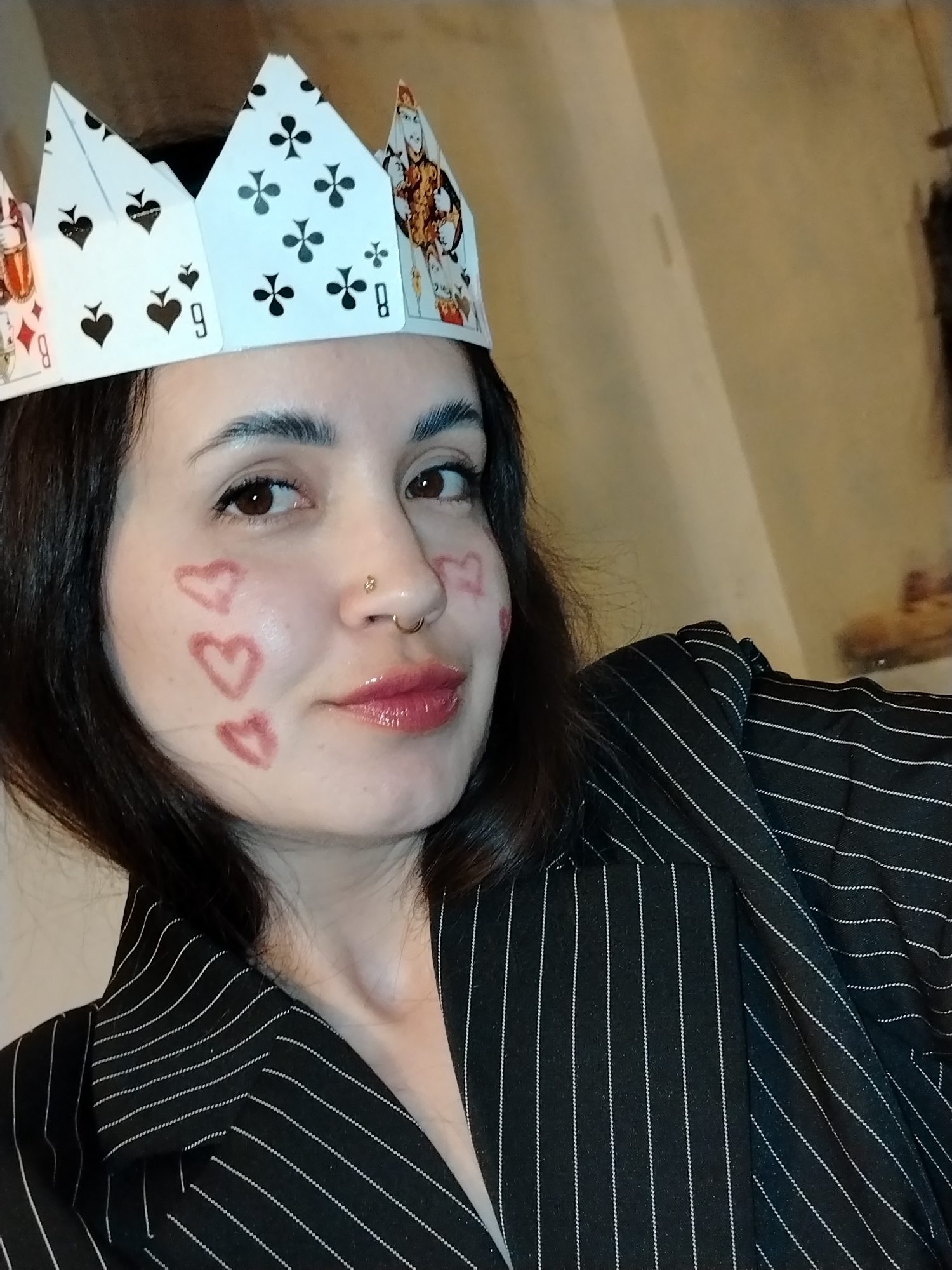 A young woman is featured in a close-up indoor portrait, looking directly at the camera with a gentle, pleasant smile. She has dark hair and eyes, and her skin appears fair. She is wearing a whimsical crown made from playing cards, which prominently display suit symbols like spades and clubs, along with numbers such as "9" and "8," and a King card. Her left cheek is adorned with several hand-drawn heart shapes, seemingly done with red or pink lipstick or makeup, with one also visible on her right cheek. She sports a small golden stud piercing on her right nostril and a golden ring piercing on her left. Her outfit includes a dark jacket or blazer with fine white pinstripes. The background is a plain, light brown or beige wall, indicating an indoor setting with warm, artificial lighting. The playful crown and face art suggest a festive, themed, or costume event. No specific location cues are visible in the image itself, beyond the indoor setting, but the context is Noviy, Russia.