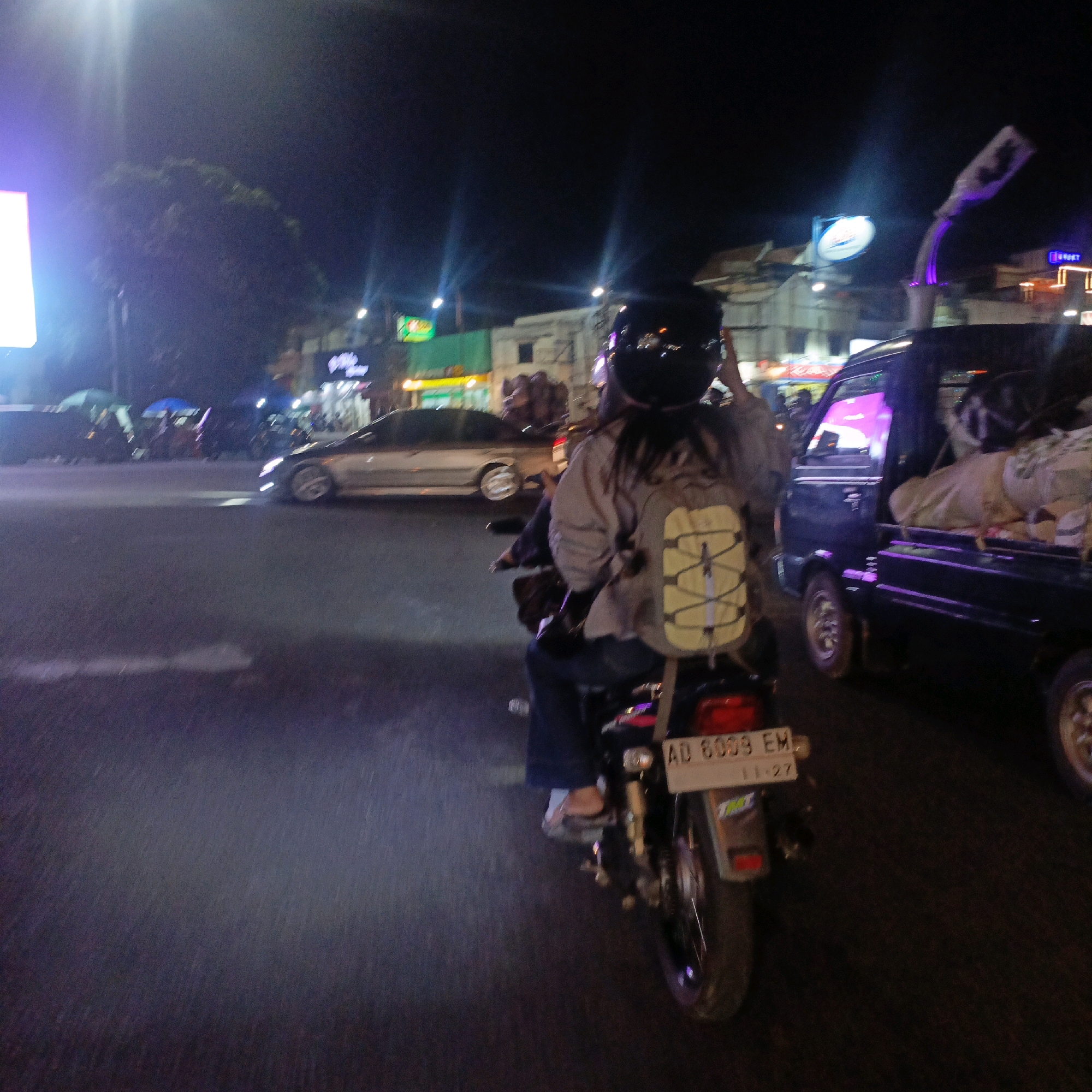 The image depicts a night scene on a street in Boyolali, Indonesia, likely in the vicinity of the city center. A person wearing a black helmet and a grey jacket with a yellow-strapped backpack is riding a motorcycle on the road. The motorcycle has a license plate that reads "AD 6009 EM" and a smaller sticker below it. To the right of the motorcycle, a dark blue pickup truck is visible, loaded with what appears to be white sacks or fabric. In the background, various buildings and streetlights are illuminated, suggesting a commercial or urban area. A silver car is also visible driving in the opposite direction. The road surface is asphalt, and there's a slight blur suggesting movement. The lighting conditions indicate it is nighttime, with artificial lights from buildings and vehicles creating reflections and highlights.