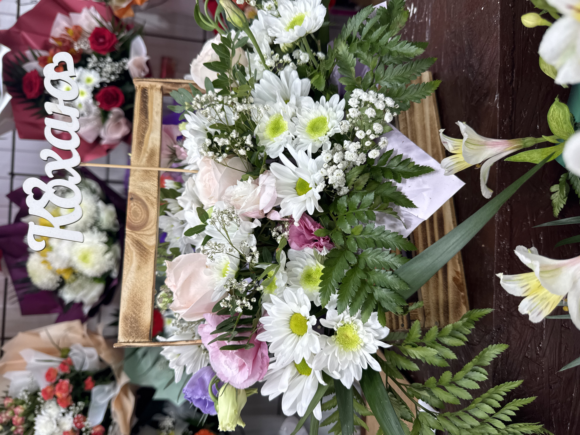 The image shows a close-up view of a floral arrangement displayed in a wooden basket. The arrangement is predominantly white and pink, with accents of purple and green foliage. Several white daisies with bright yellow centers are prominent, along with delicate pink roses and sprigs of baby's breath. A wooden sign with white lettering, appearing to be Cyrillic characters, is positioned at the top of the arrangement. The background is blurred, showing other bouquets of flowers, suggesting the setting is a flower shop or market. The image appears to be taken in Kamianske, Ukraine, given the context provided.
