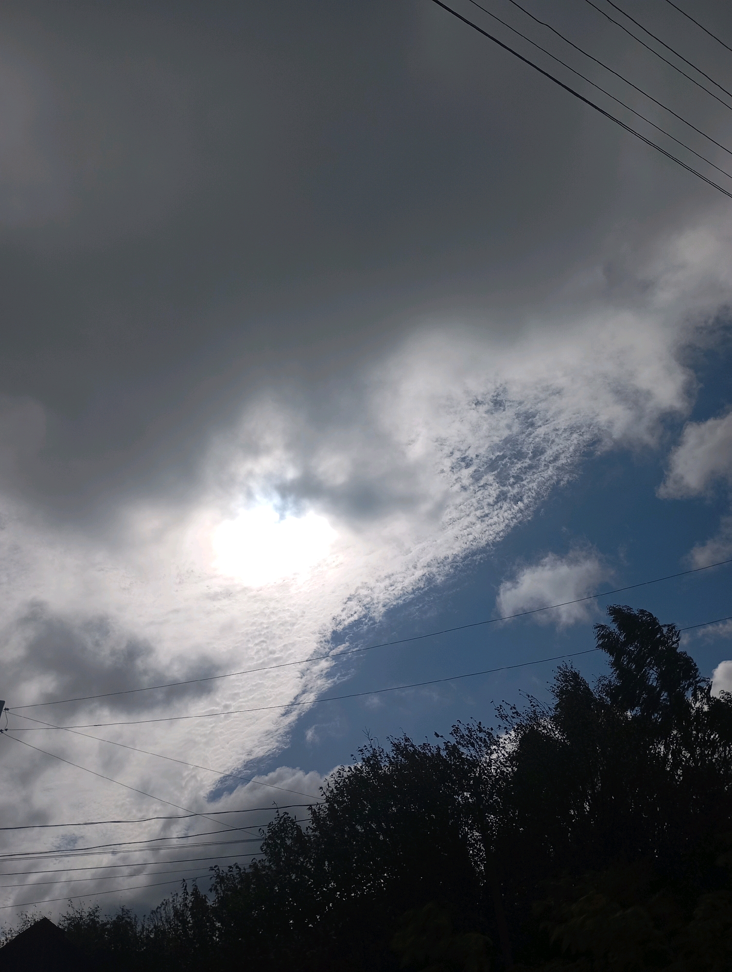 The image depicts a cloudy sky with the sun partially visible through a gap in the clouds. Several power lines are visible running diagonally across the frame from the top right to the bottom left. The lower portion of the image is dominated by the dark silhouettes of trees and a building or roofline. The scene is likely an outdoor environment in or near the city of Fokinskoye, Russia.