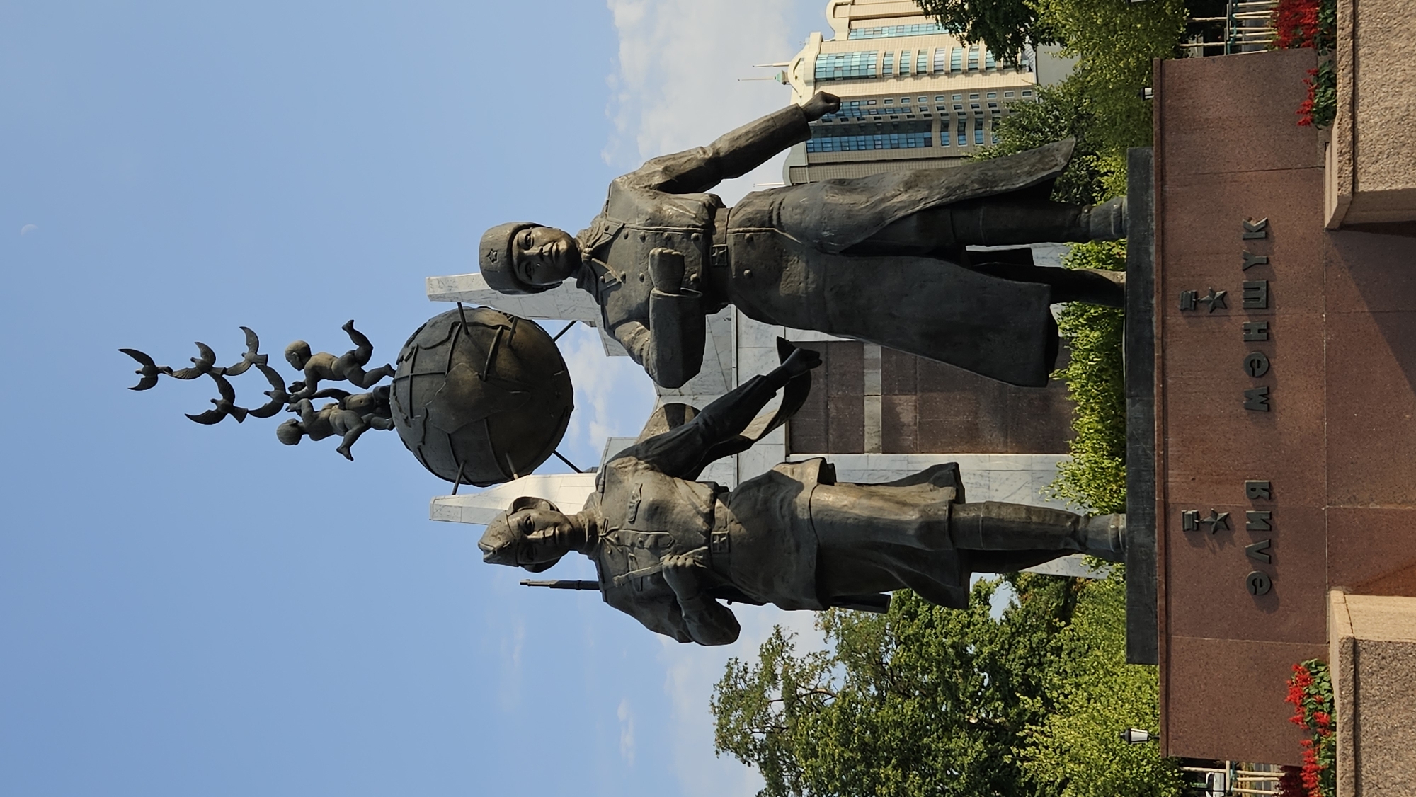 This is a full-shot, eye-level, vertical photograph of the "Eternal Flame" memorial in Almaty, Kazakhstan. The memorial features two bronze statues of women in military uniforms standing in front of a large globe supported by a marble structure. Above the globe, a smaller sculpture depicts children reaching for a flock of birds. The base of the memorial has inscriptions in Cyrillic script. In the background, there are green trees, a modern high-rise building, and a clear blue sky with a crescent moon visible. The foreground shows part of the memorial's granite base with red flowers planted in front of it.