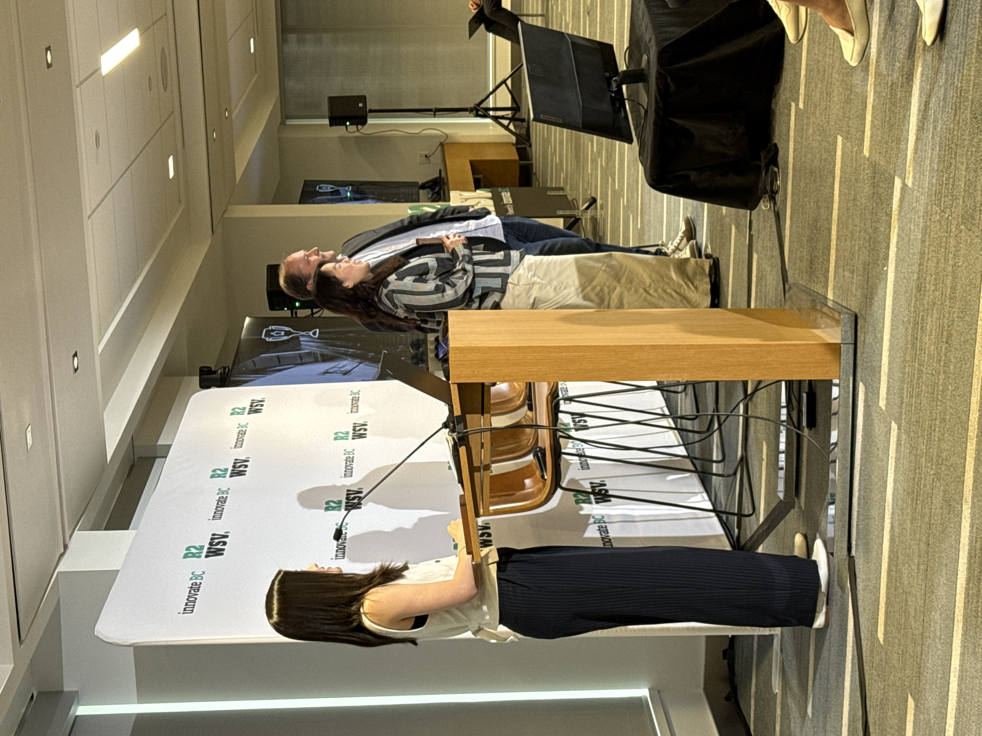 Here's a detailed description of the image:
**Scene:** The image appears to be taken at an event or conference, potentially related to innovation or business in British Columbia. The backdrop features the words "innovate BC", "R2" and "WSV" in green text.
**People:**
* A person, most likely a woman is standing behind a wooden lectern, facing the backdrop. She has dark hair and is wearing a light-colored top and dark pants.
* A man and woman are standing behind the lecturn in the background.
**Objects:**
* Wooden lectern with microphone attachments
* Backdrop with branding
* A screen or projector with an image displayed on it in the background
* Chairs
**Environment:** The event seems to be taking place in an indoor venue, possibly a conference hall. The lighting is bright.
