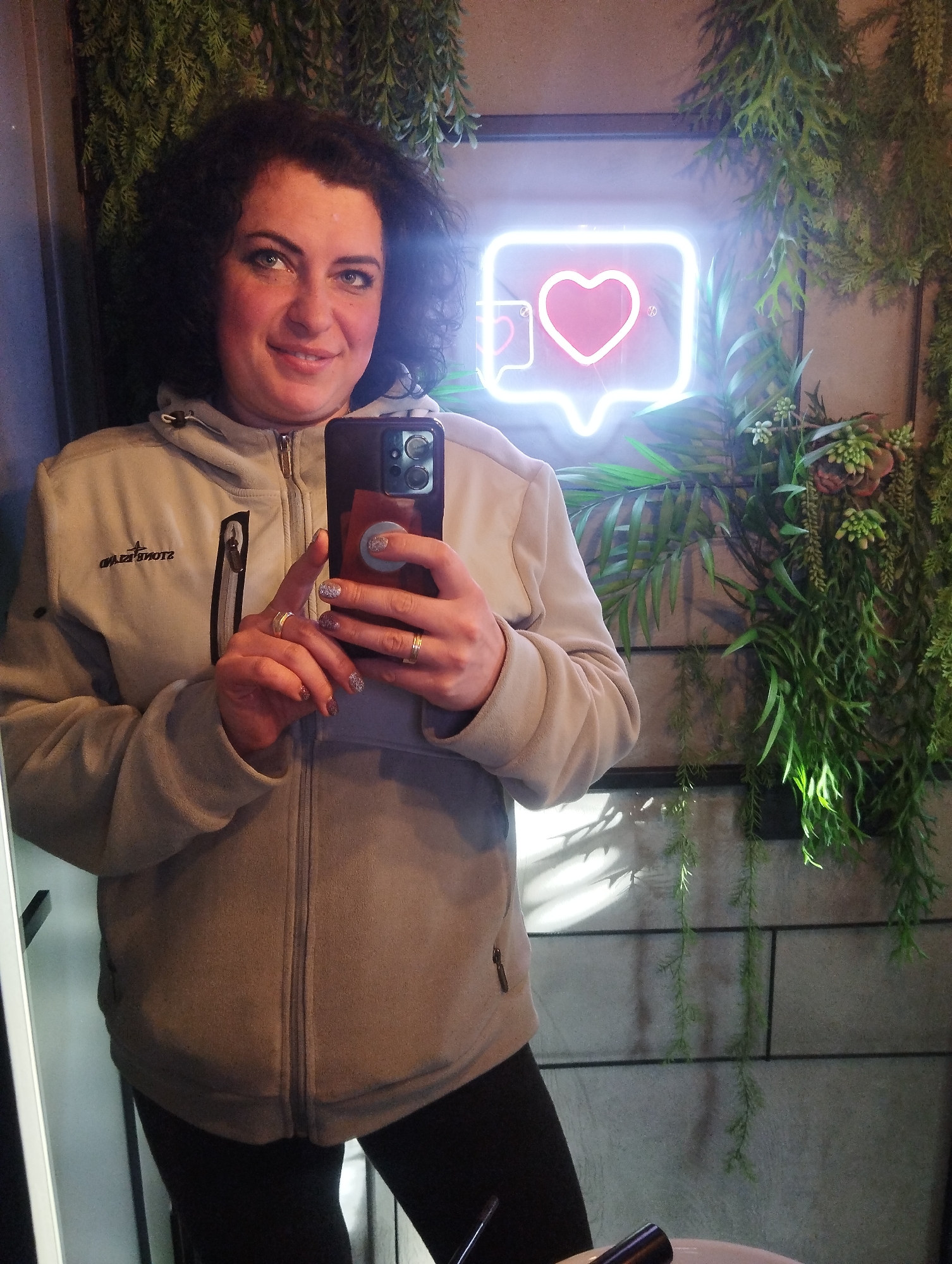 A woman with dark curly hair is taking a selfie in what appears to be a bathroom. She is wearing a light-colored fleece jacket with a hood and black leggings. The background features a wall decorated with faux plants and a neon sign in the shape of a social media notification with a heart inside. The bathroom has tiled walls. There is no contextual evidence to confirm the location in the city of Votkinsk, Russia.