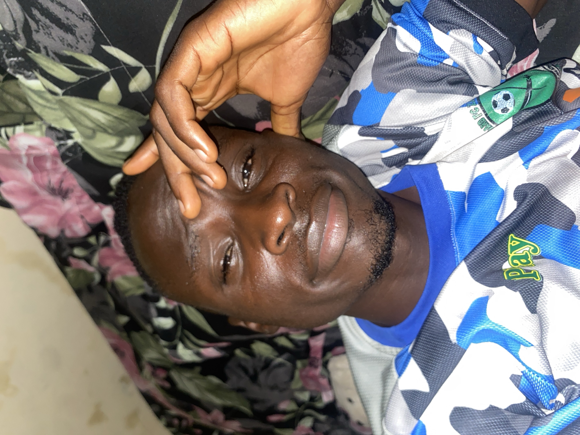 A close-up, downward-facing shot shows a person's face and chest, angled to the right. The person, whose skin tone is dark, appears to be lying down on a patterned surface, possibly a bedspread, with floral motifs in pink and green against a dark background. Their right hand rests on their forehead and temple, with fingers slightly spread. They are wearing a short-sleeved shirt with a camouflage pattern in shades of blue, gray, and white. A green emblem with a soccer ball and text in white and green is visible on the left side of the shirt. The person's left eye is visible, looking slightly to the left, with a small beard visible on their chin and jawline. The overall lighting suggests an indoor setting. The image does not provide specific visual information about the city of Barden Yana, Nigeria.